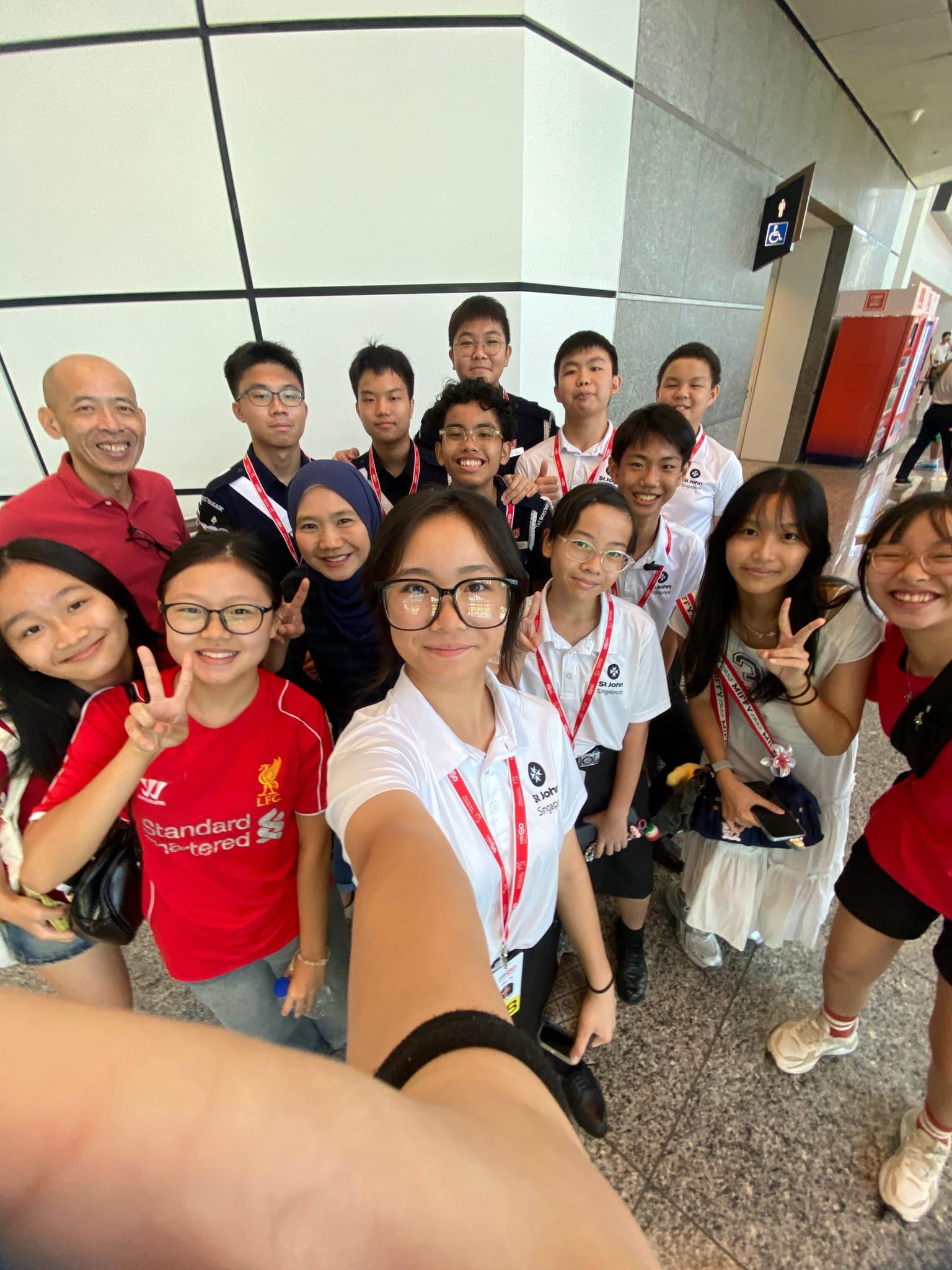 A group of smiling teenagers and adults pose for a selfie indoors, several wearing red lanyards and white St John uniforms.