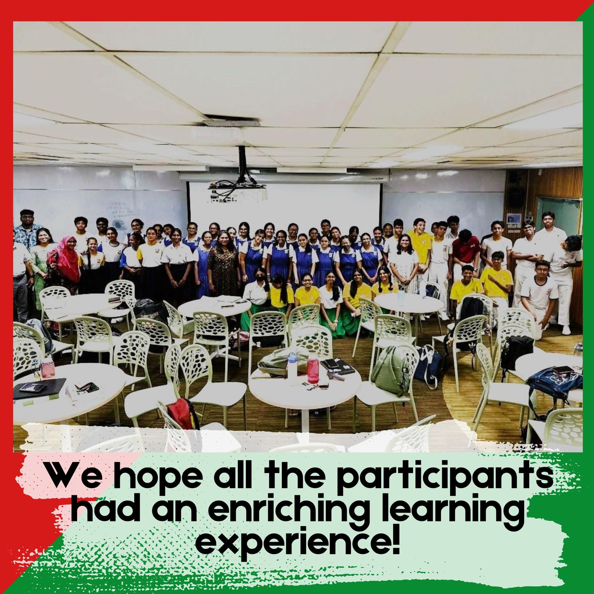 A large group of students in uniforms posing together in a classroom with round tables and a projector screen.