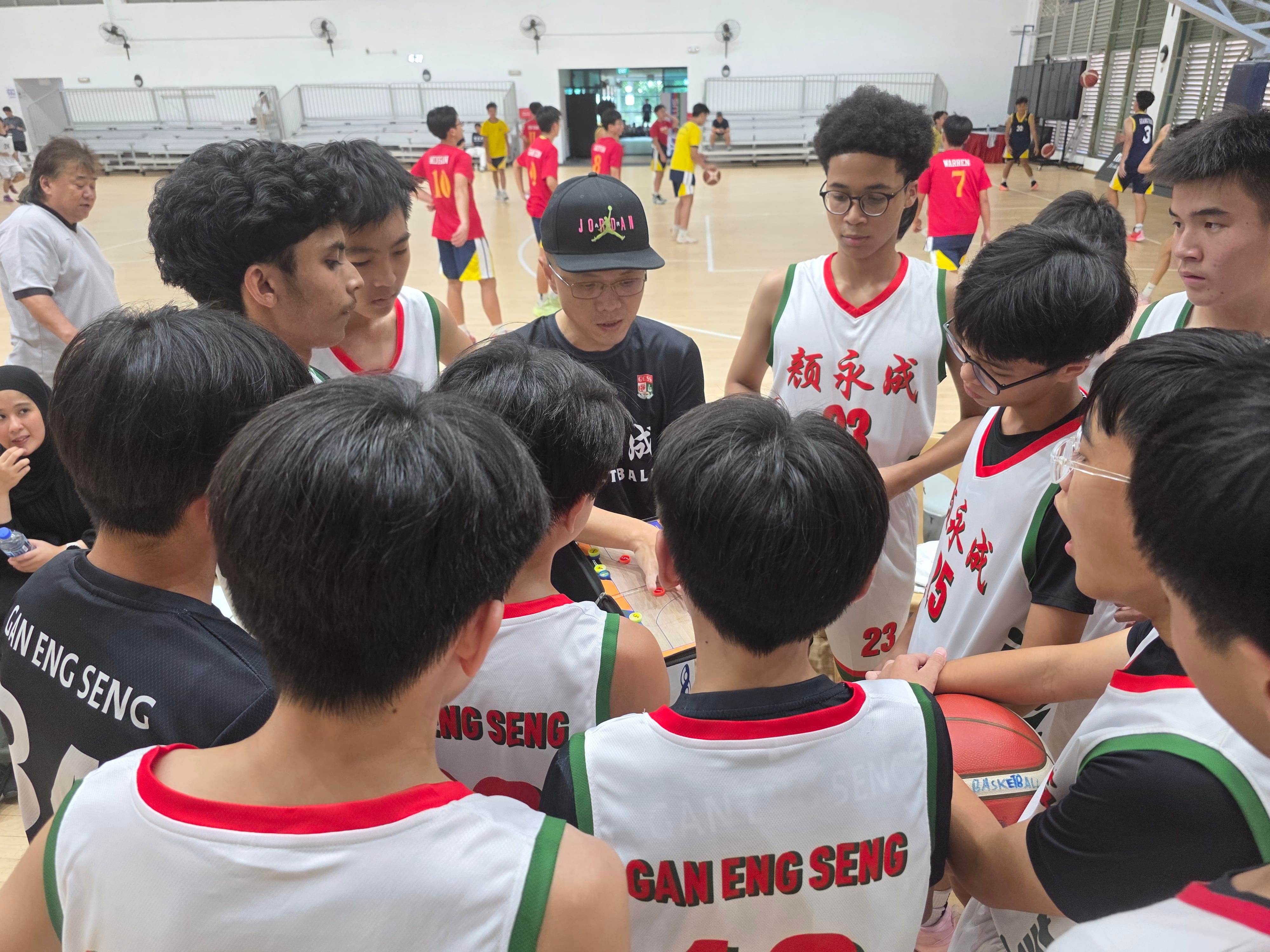 A coach huddles with Gan Eng Seng basketball players in white jerseys, reviewing a play board inside an indoor court.