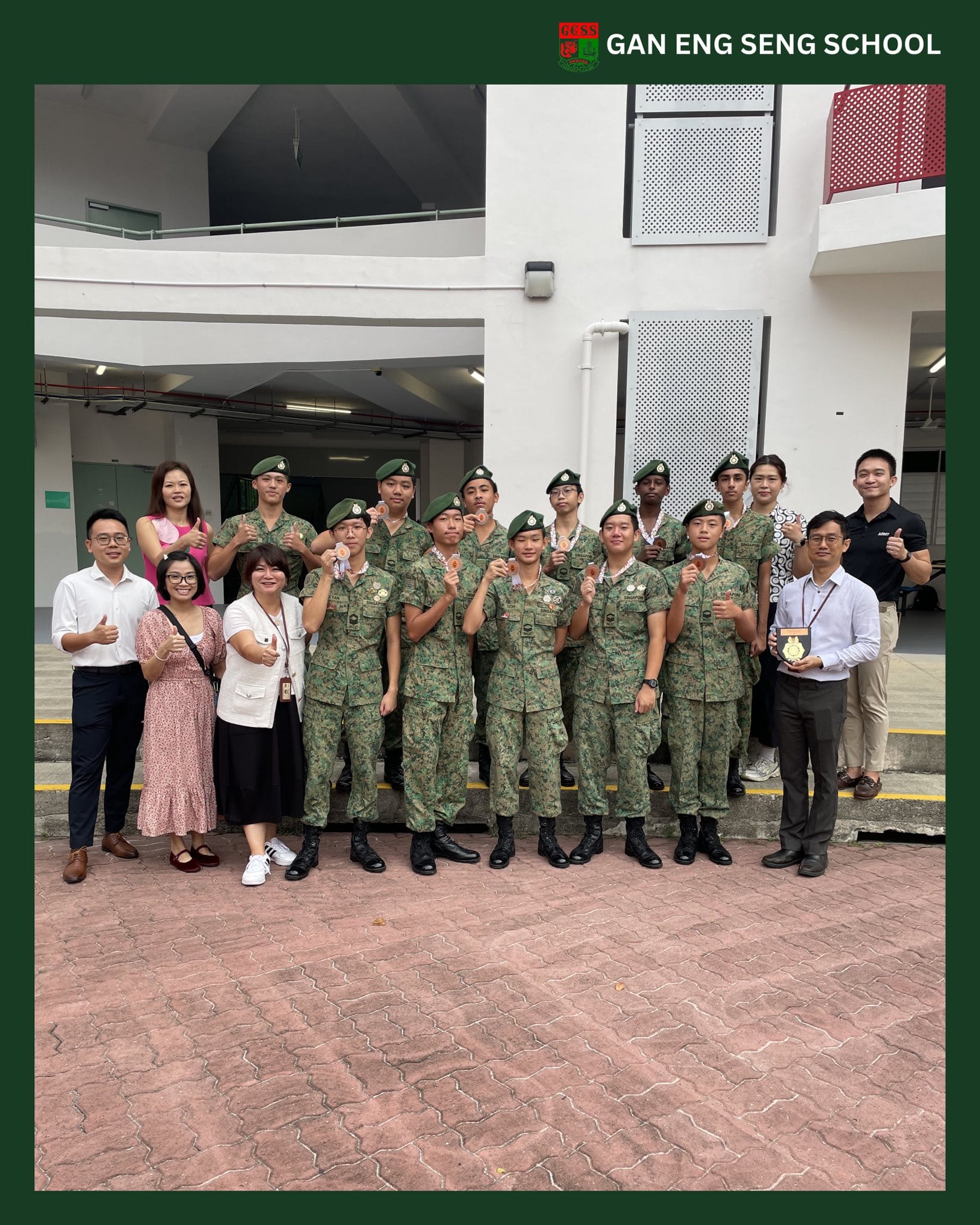 Group of students in green camouflage uniforms holding medals, posing with teachers outside Gan Eng Seng School.