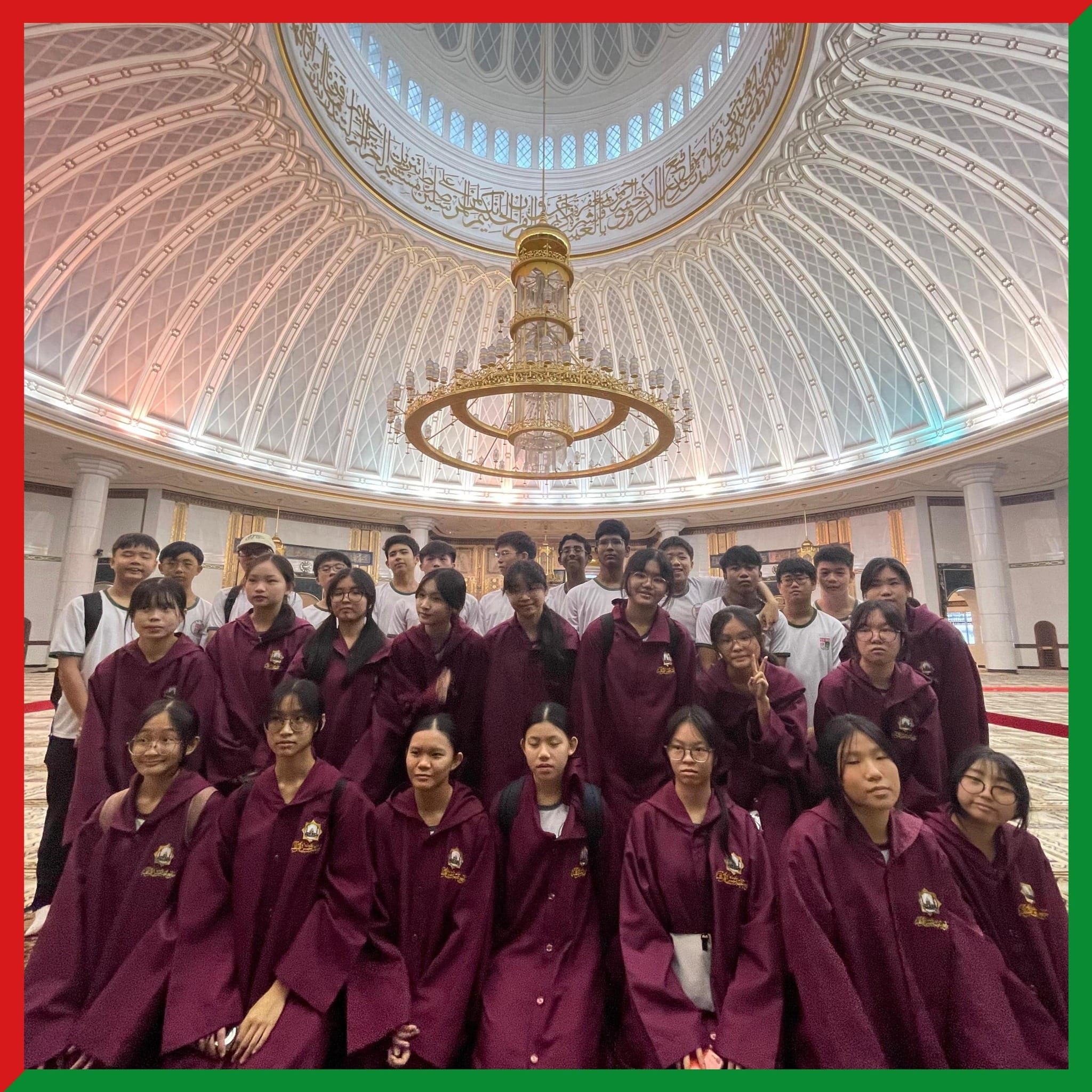 A group of students in maroon robes pose inside a grand mosque with a large ornate dome and gold chandelier.