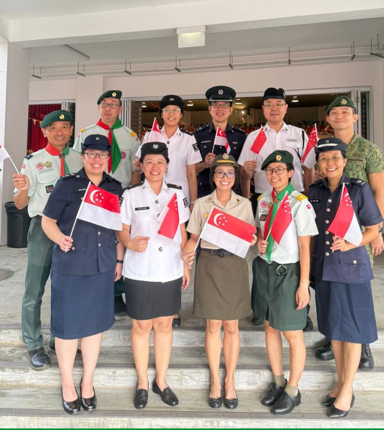 Group of uniformed people posing on steps, holding small Singapore flags and smiling at the camera.