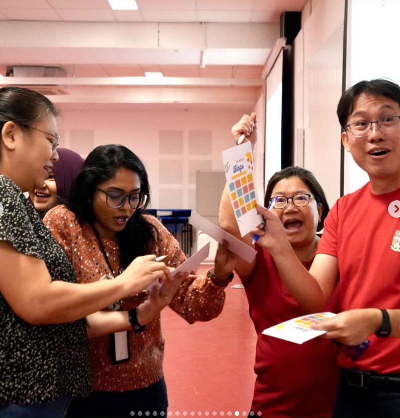 Four people laughing and reacting excitedly while holding colorful bingo cards in an indoor room.