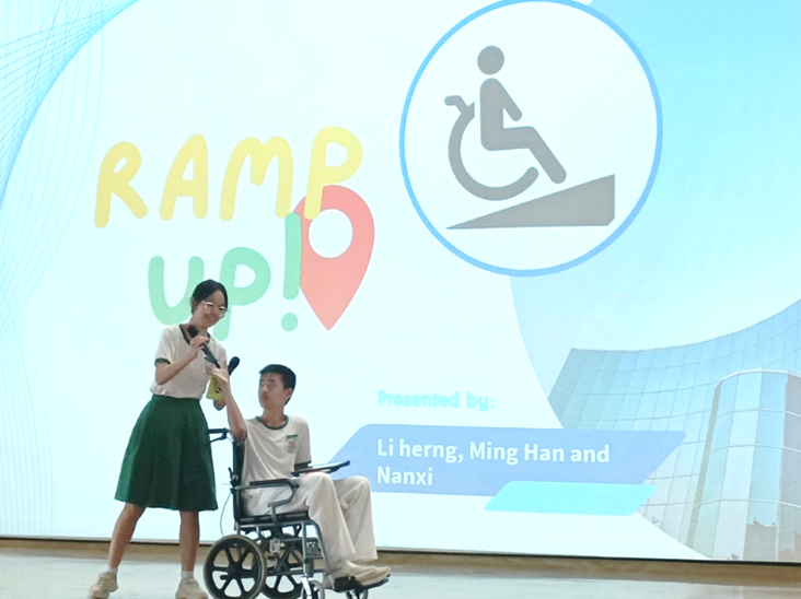 A girl holds a microphone while a boy sits in a wheelchair during a presentation titled Ramp Up on a projected screen.