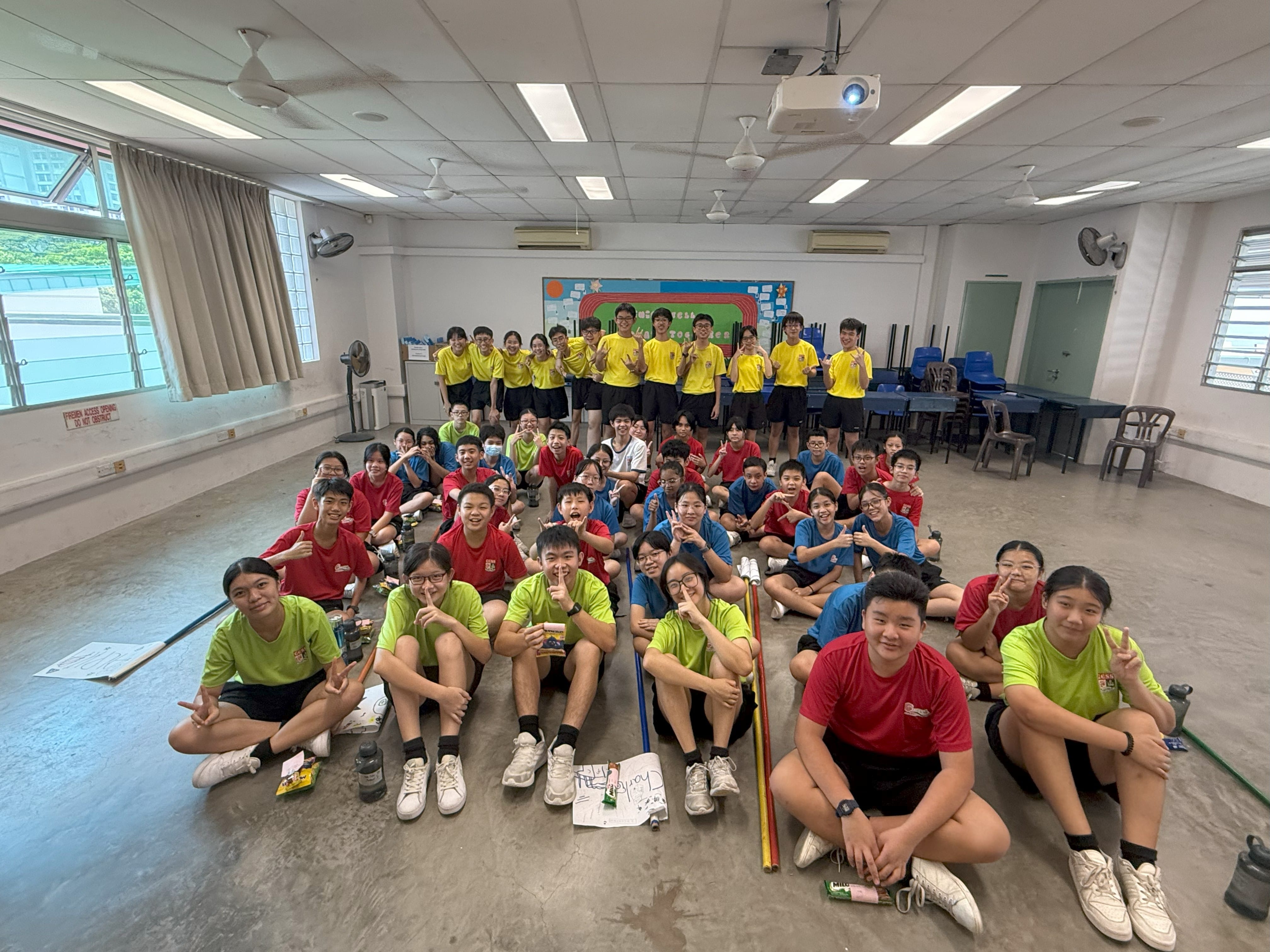 A large group of students in yellow, red, blue, and green shirts pose together in a school hall.