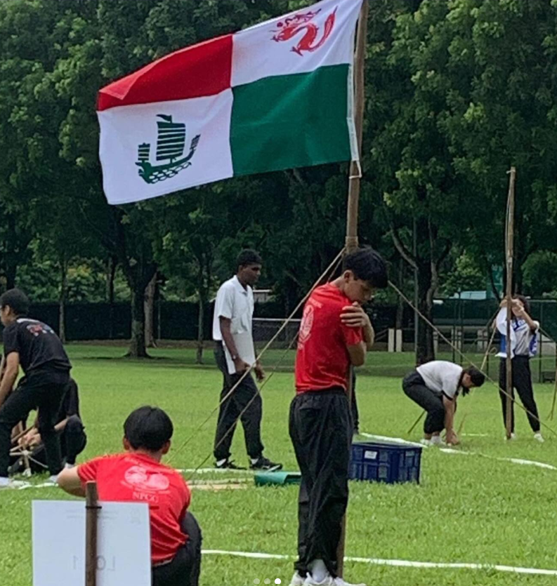 Several students on a grass field erect wooden poles with a red, white, and green flag flying above them.