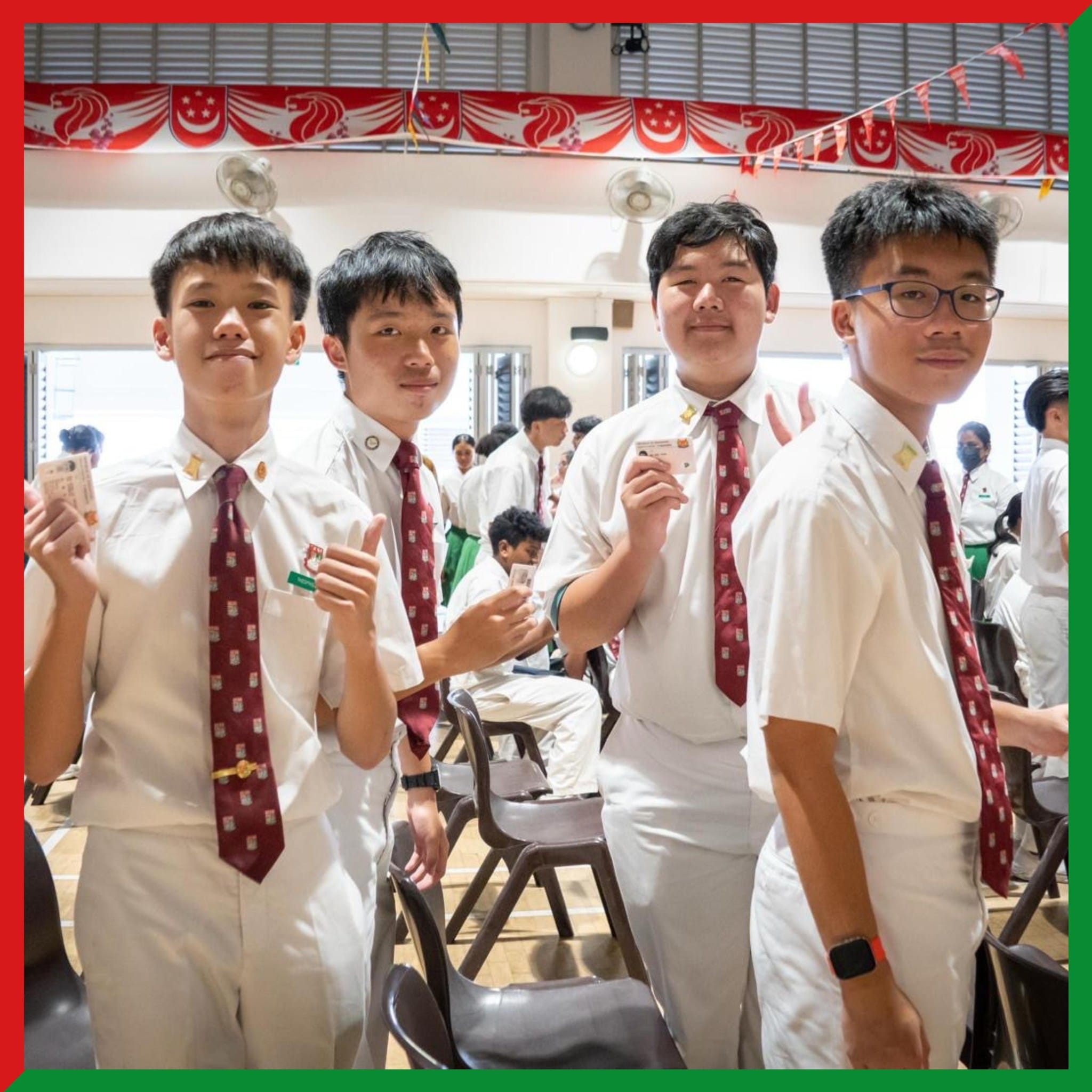 Four male students in white uniforms with dark red ties smile and hold up cards in a decorated school hall.