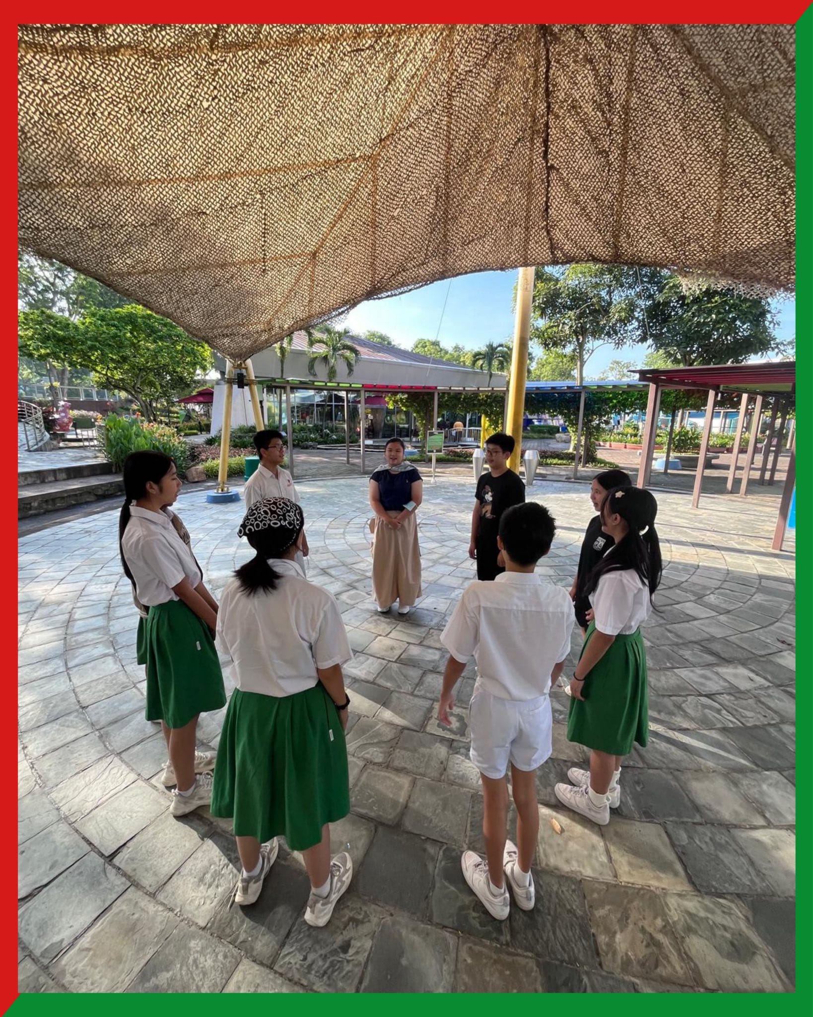 Students in white shirts and green skirts stand in a circle on a paved outdoor plaza under a woven shade canopy.