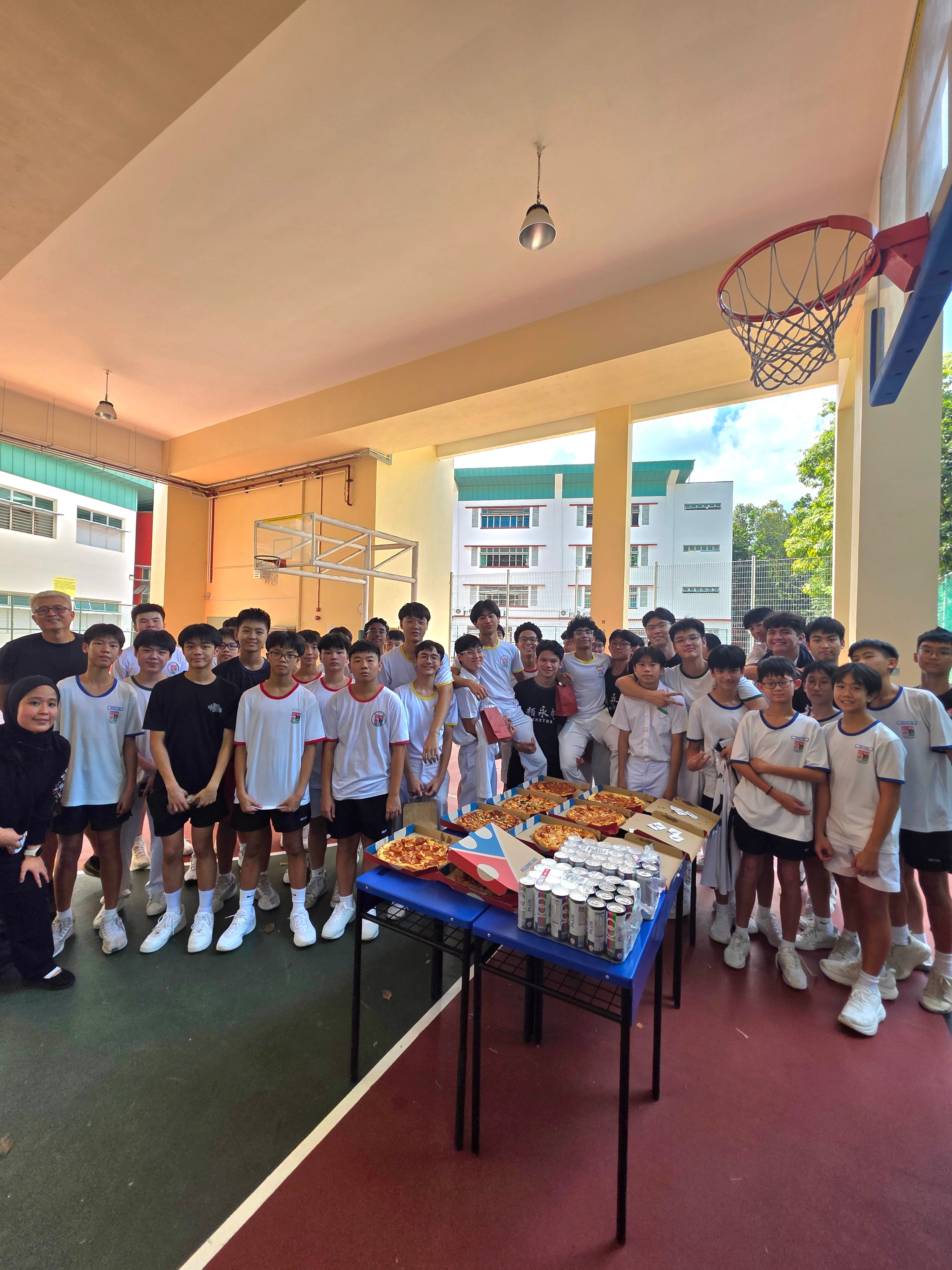 A large group of students in white uniforms pose on a school basketball court beside a table with pizzas and canned drinks.
