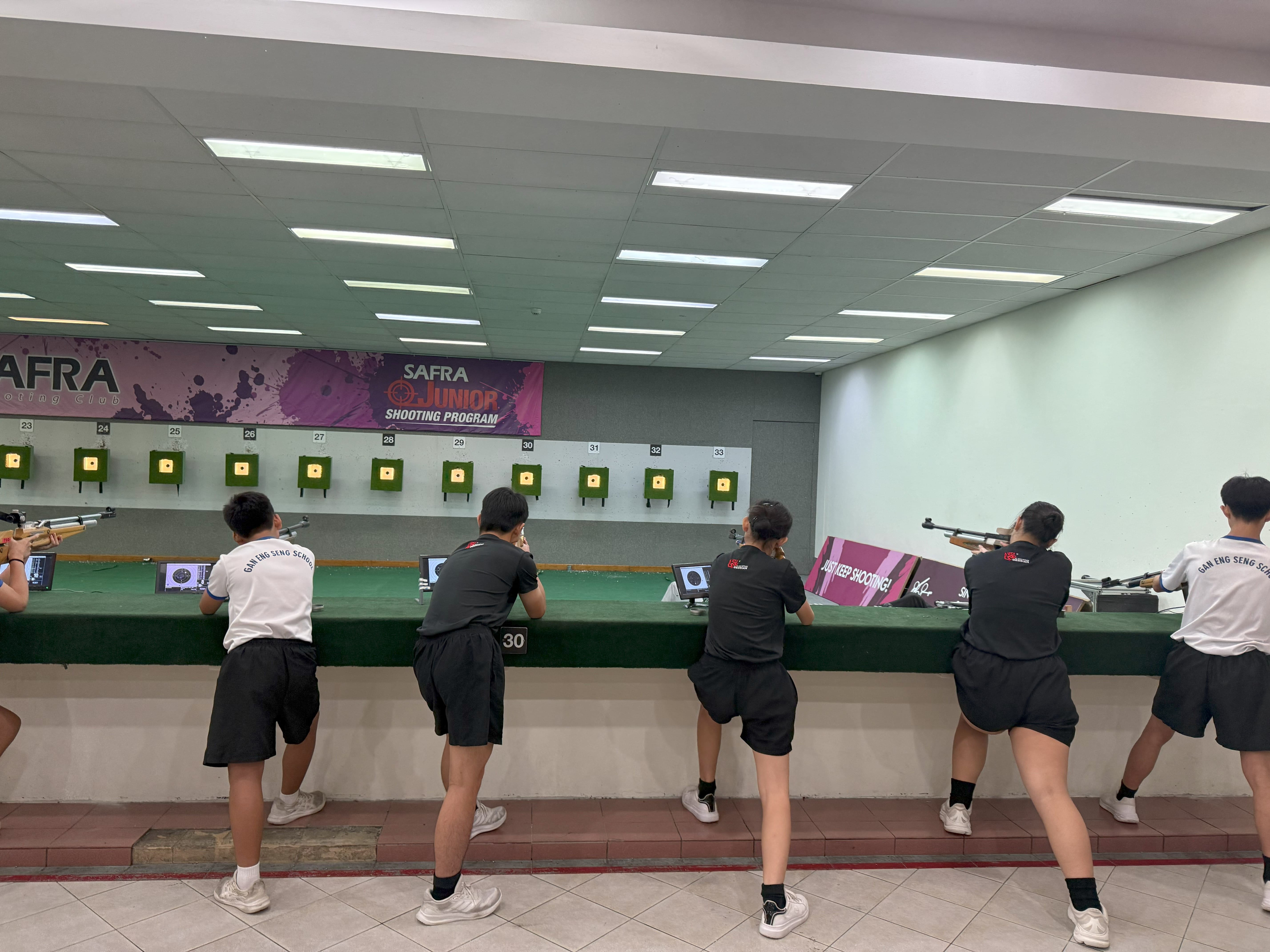 Several young students aim rifles at targets in an indoor SAFRA Junior Shooting Program range.