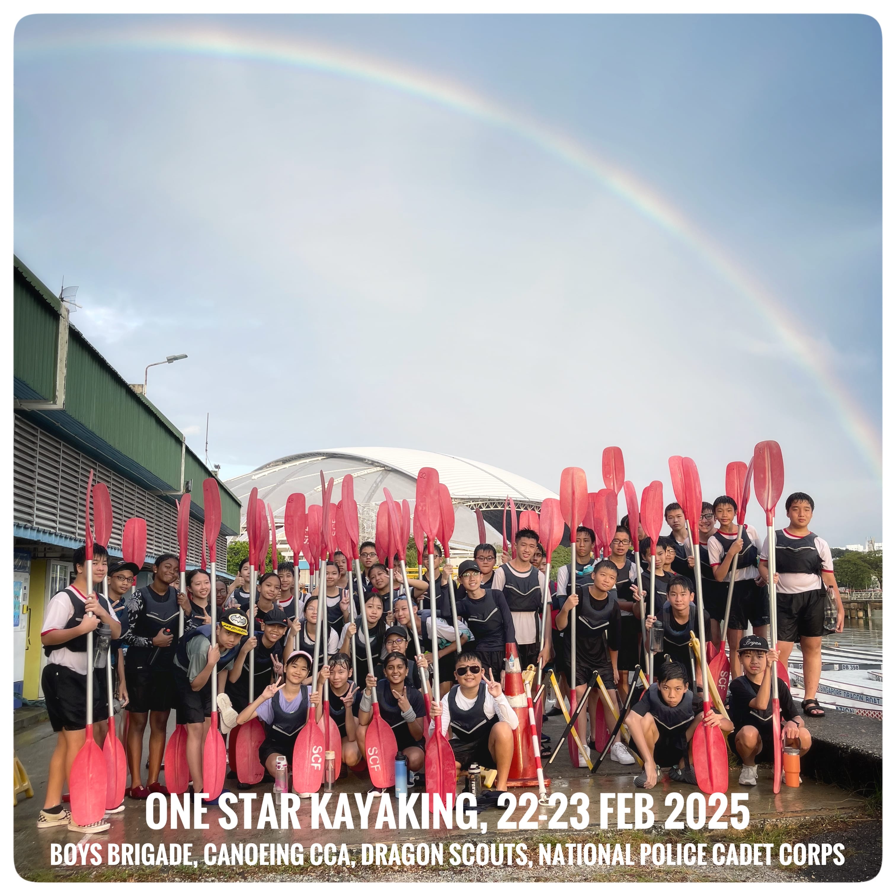 Large group of youths holding red kayak paddles outdoors under a rainbow, text reads One Star Kayaking 22-23 Feb 2025.