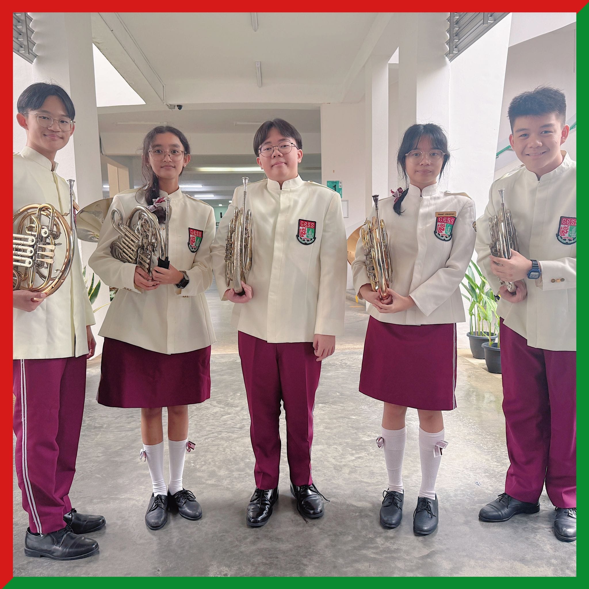 Five students in white and maroon school band uniforms hold brass instruments including French horns and trumpets indoors.