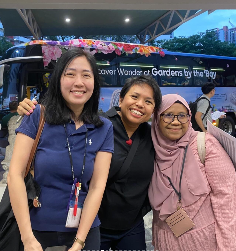 Three smiling women pose together in front of a bus with a Gardens by the Bay advertisement.
