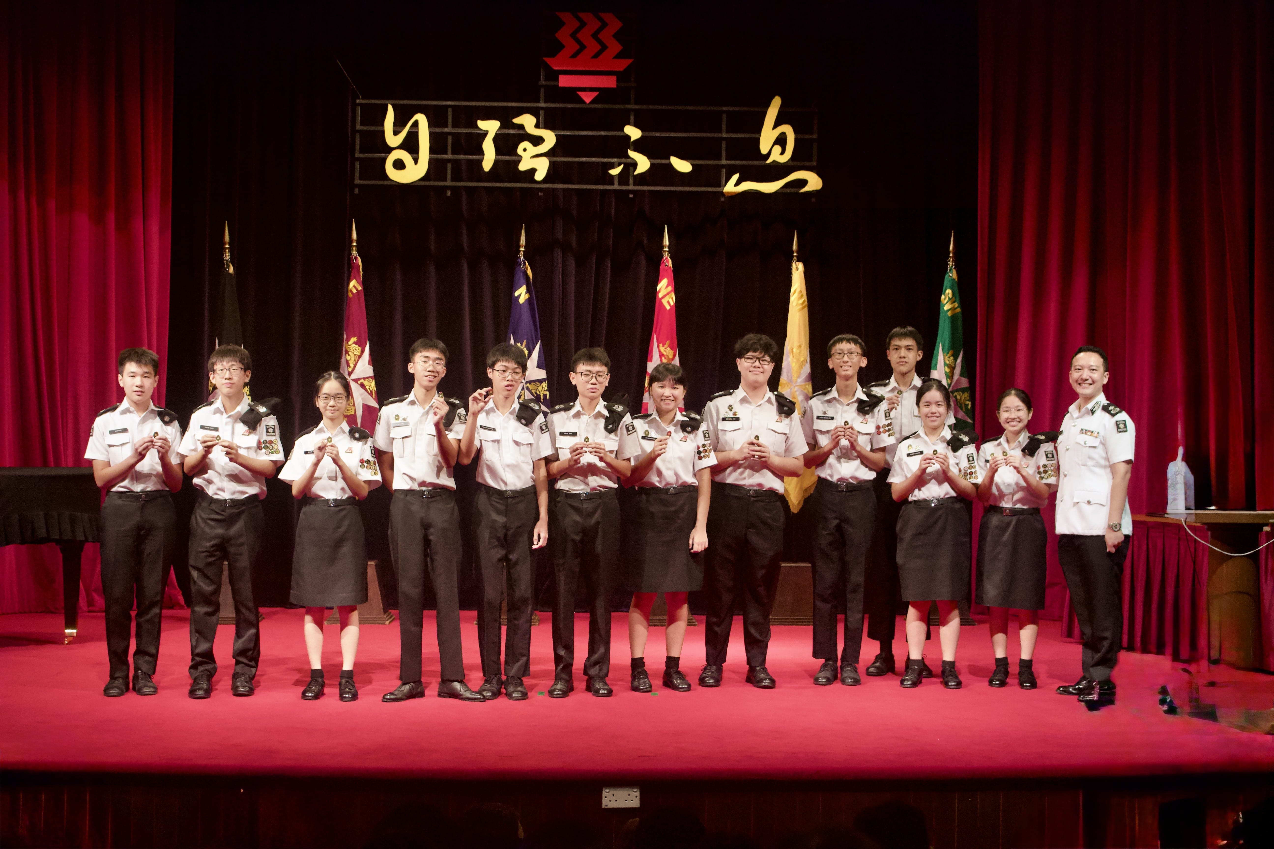 A group of young people in white and black uniforms stand on a red stage holding items, with flags and Chinese text behind them.