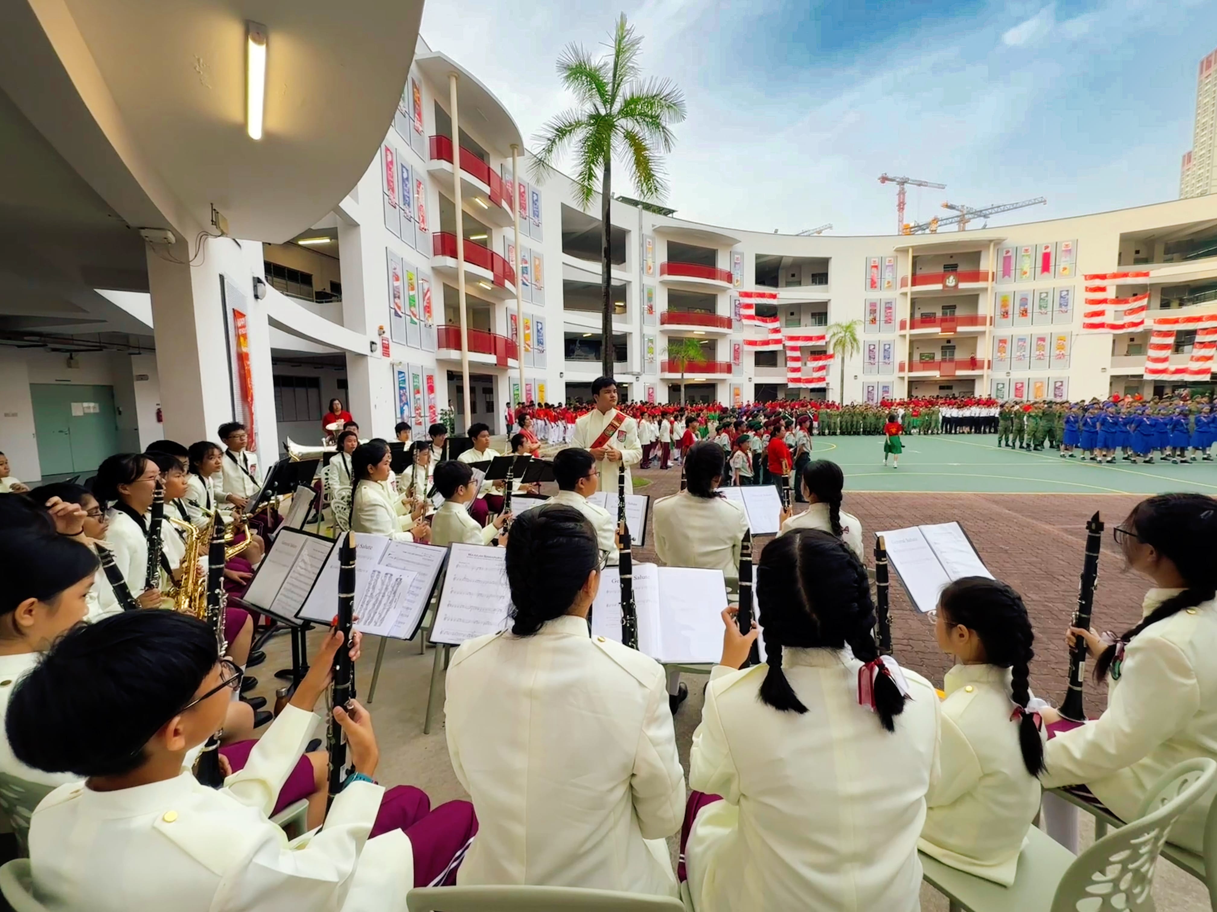 Students in white uniforms play clarinets and saxophones at an outdoor school courtyard ceremony with marching groups.