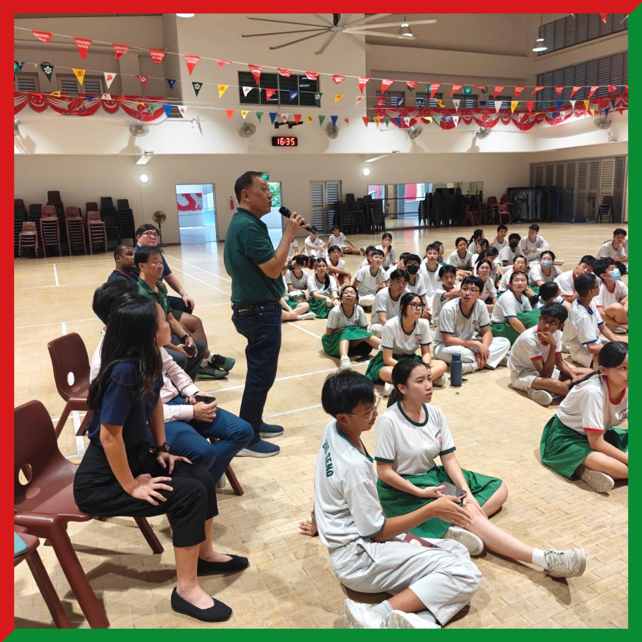 A man speaks into a microphone in a school hall while uniformed students sit on the floor listening.