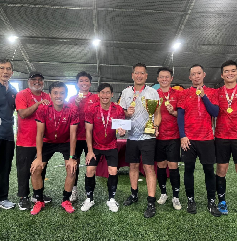 A group of men in red jerseys pose on a football pitch holding gold medals and a trophy.