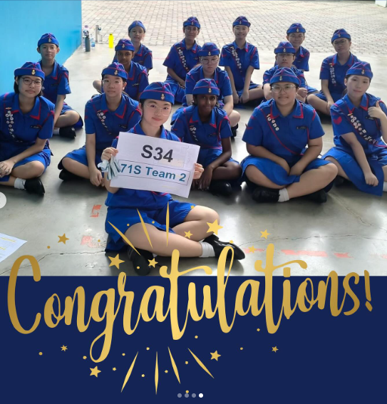 Group of youth in blue uniforms and caps sitting outdoors, one holding a sign reading S34 71S Team 2, with Congratulations text below.