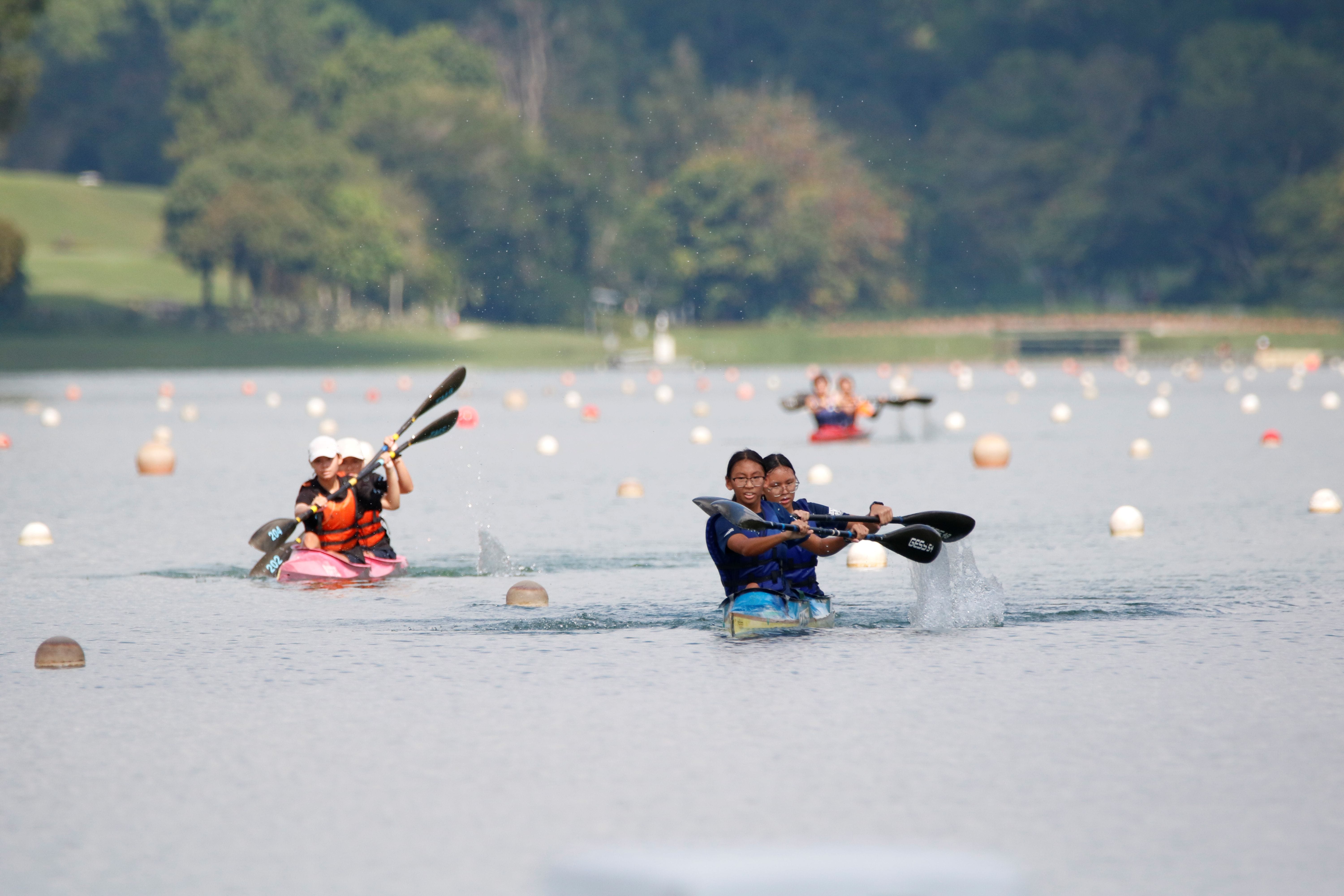 Two kayak teams paddle on a buoy-marked lake with green trees in the background.