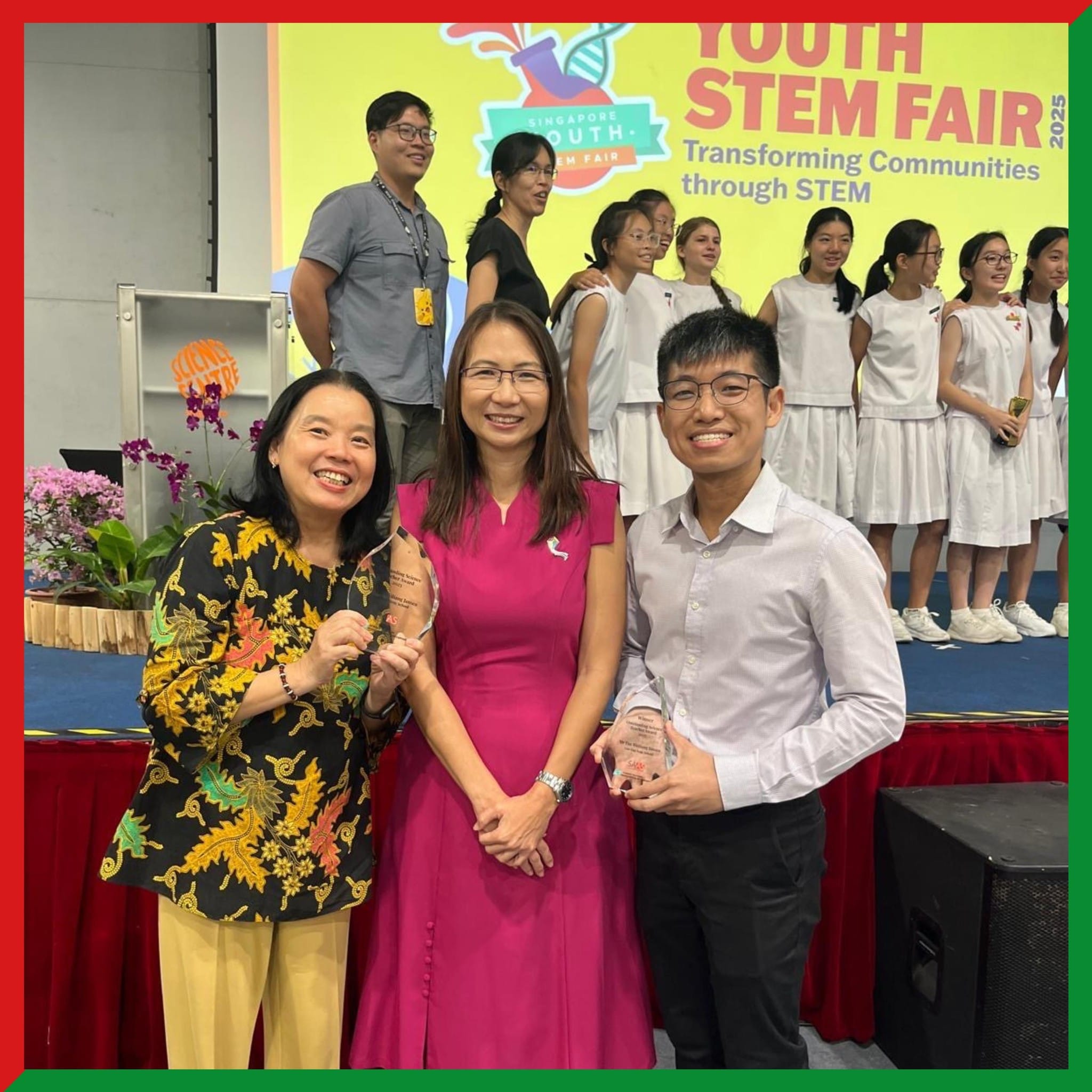 Three smiling people hold awards at the Singapore Youth STEM Fair 2025, with students in white uniforms on stage behind them.