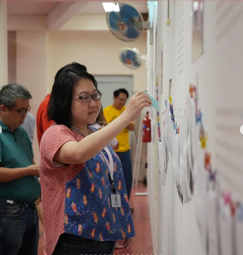 A young woman with glasses pins a paper to a wall display board in an indoor exhibition setting.