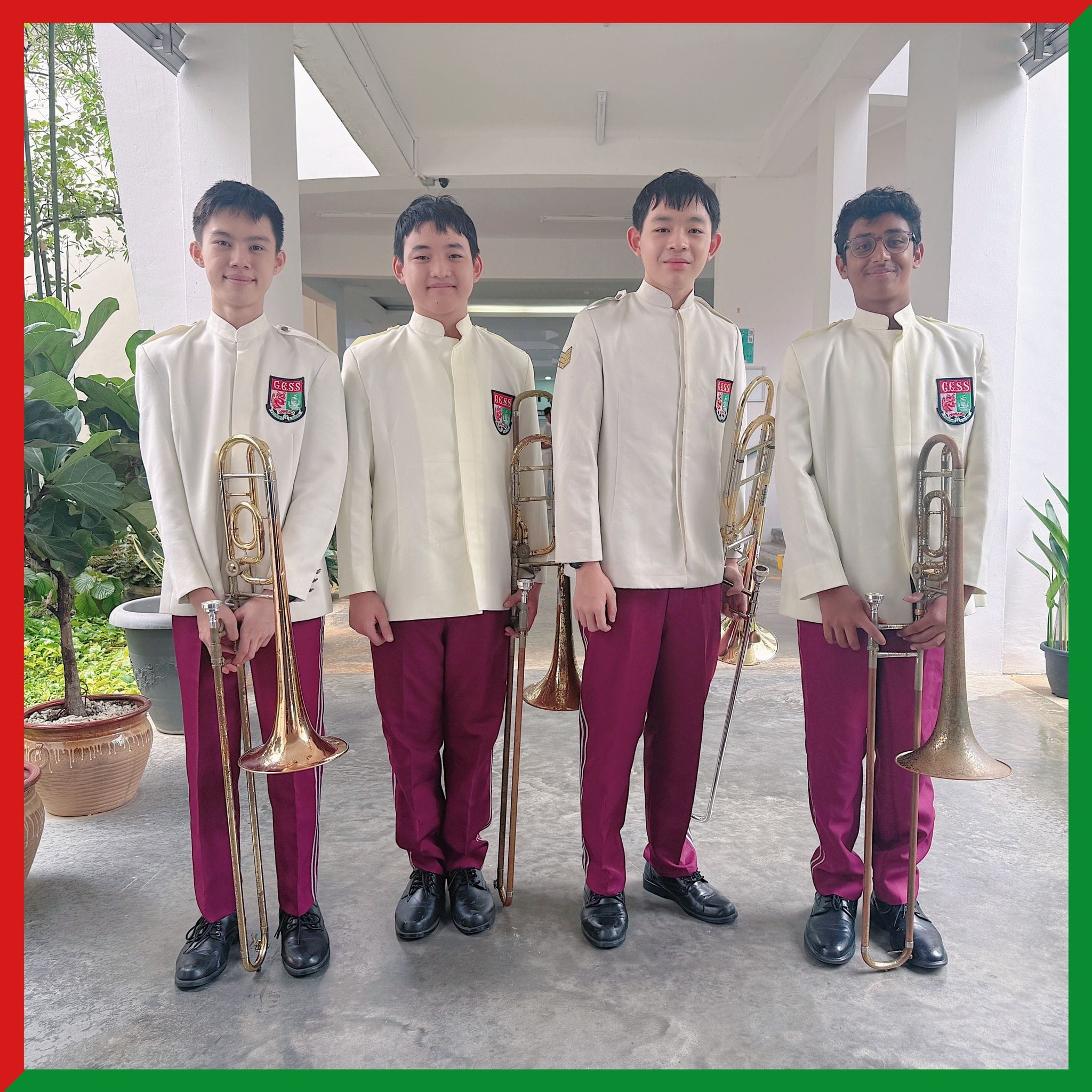 Four boys in white band uniforms with maroon pants hold trombones while standing in a school corridor.
