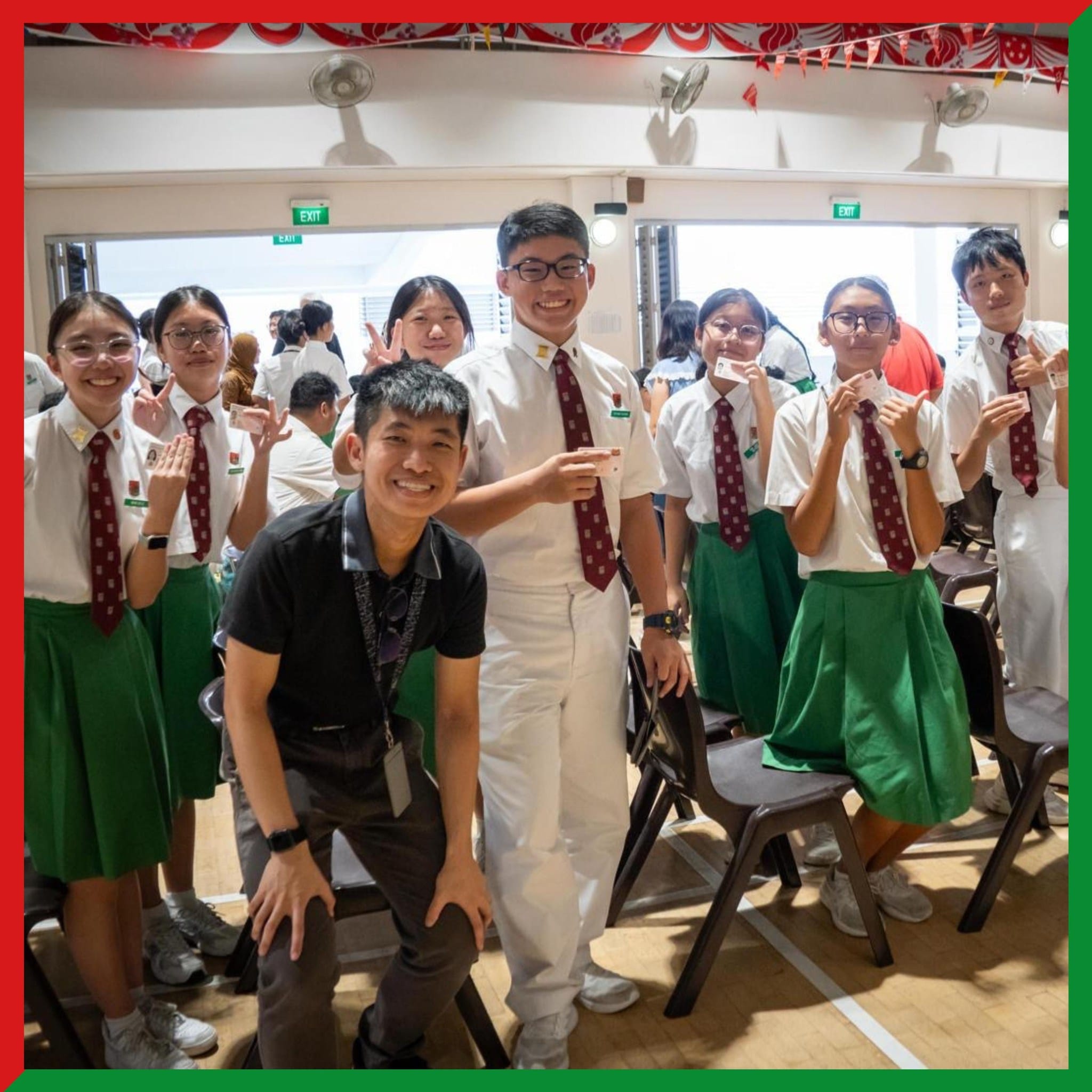 A group of smiling students in white shirts, red ties, and green skirts pose with an adult in a school hall.