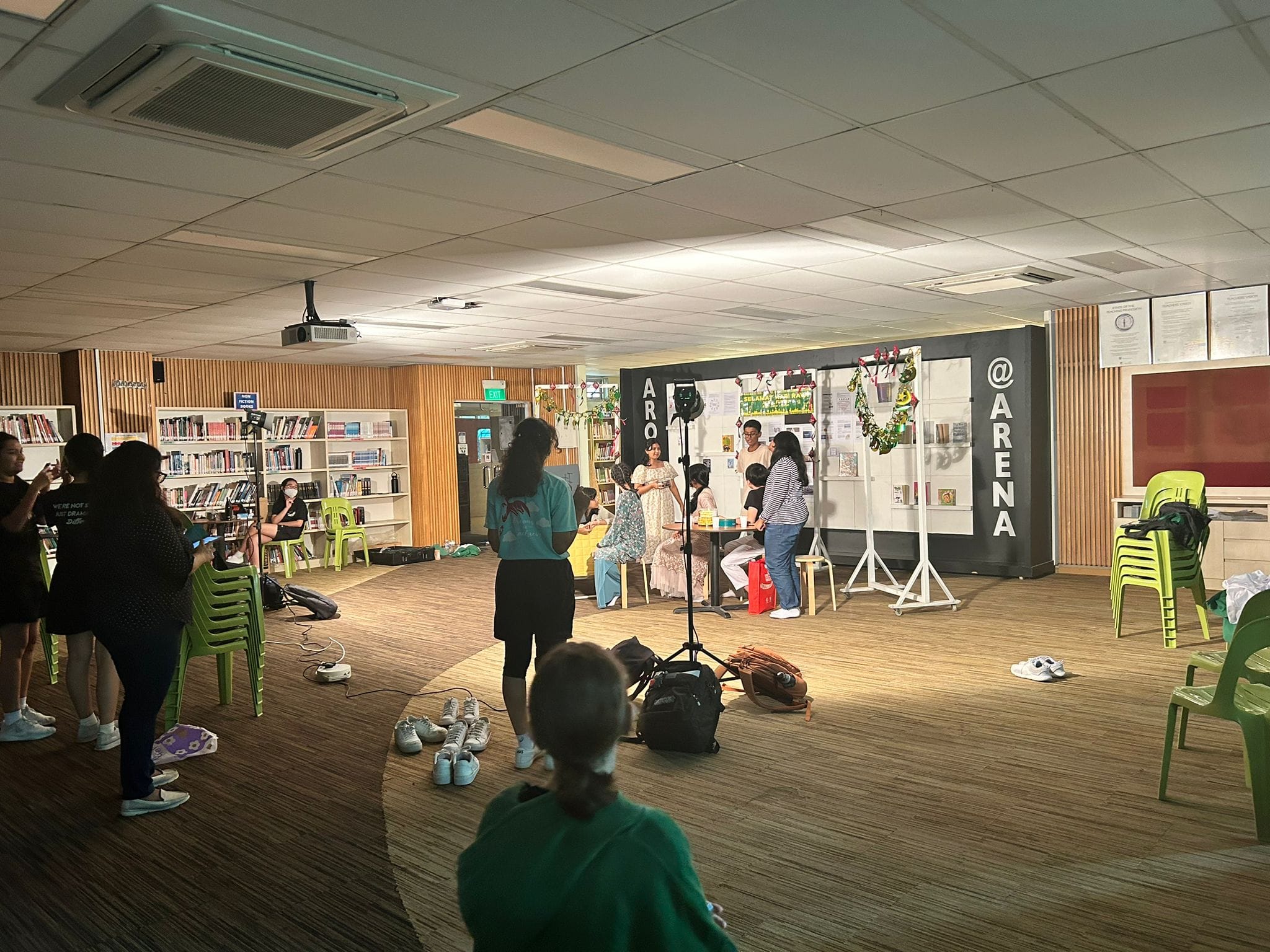 People gather in a library around a small stage with an "@ARENA" sign, with lighting equipment and green chairs nearby.