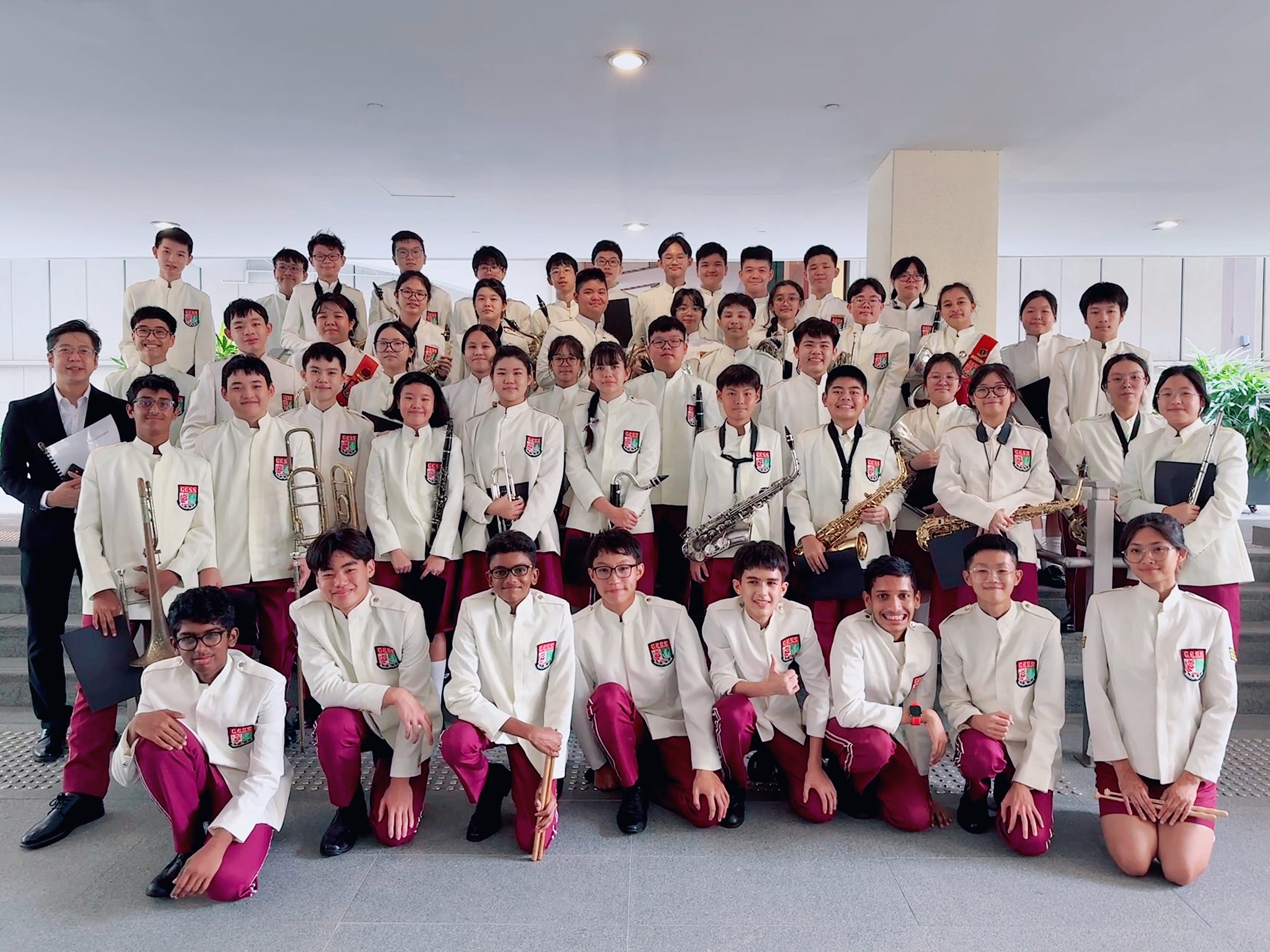 Large school band group posing in white uniforms with red trousers, holding instruments including saxophones and trumpets.