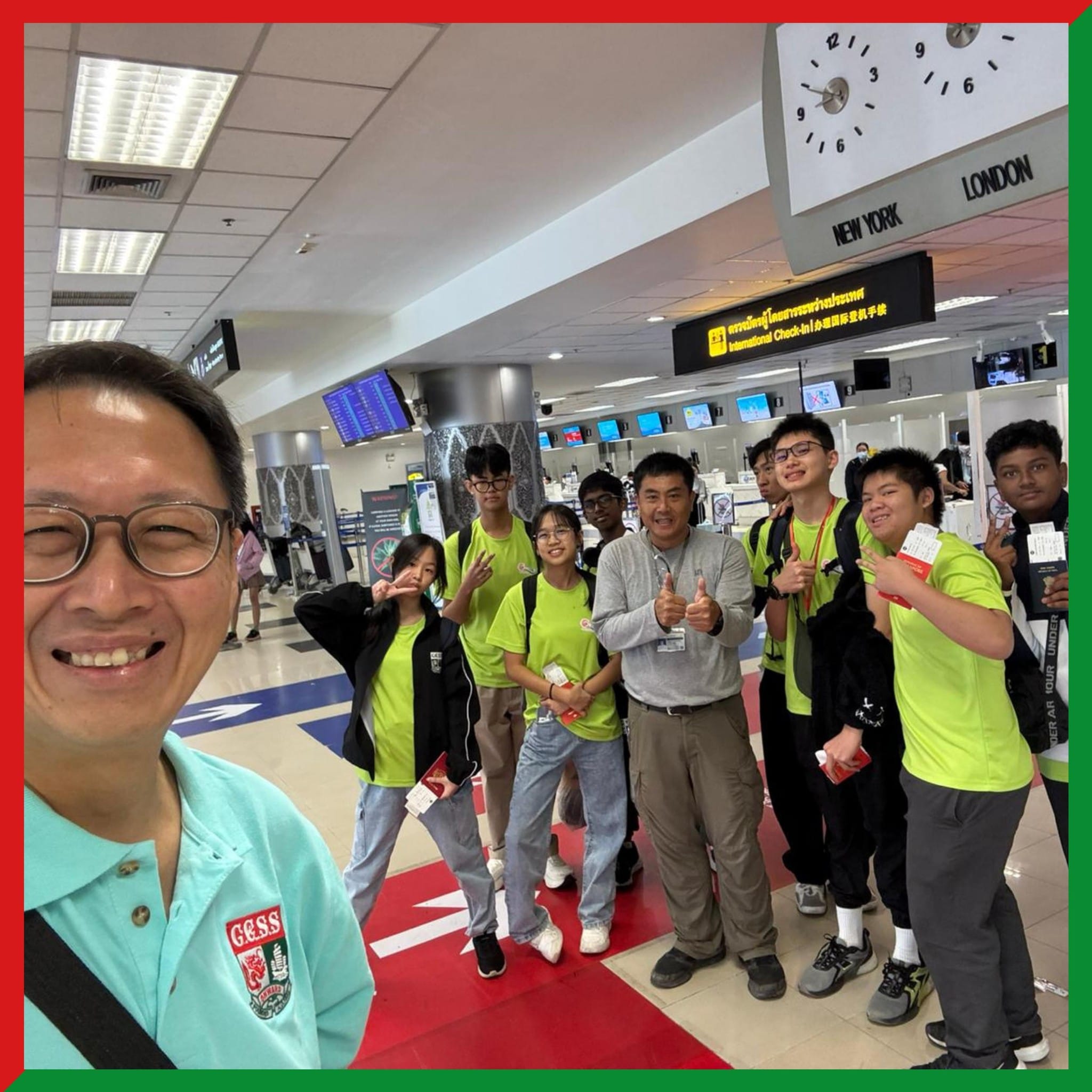 A group of students in green shirts pose with two adults at an international airport check-in area.