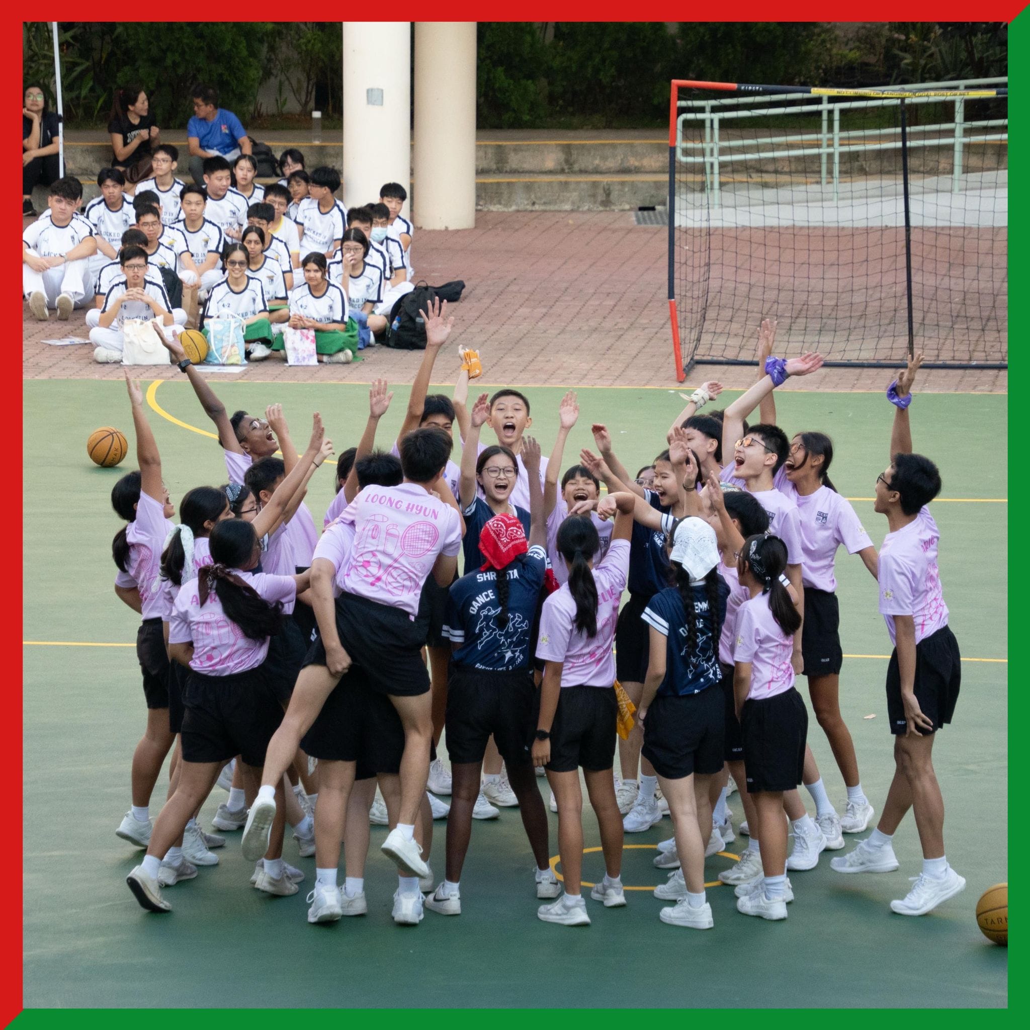 A large group of students in pink shirts celebrate with raised arms on an outdoor sports court.