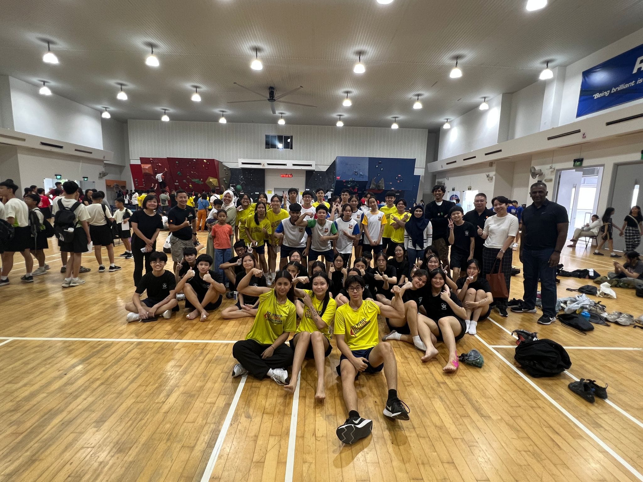 A large group of youth and adults pose together in an indoor sports hall with a climbing wall in the background.