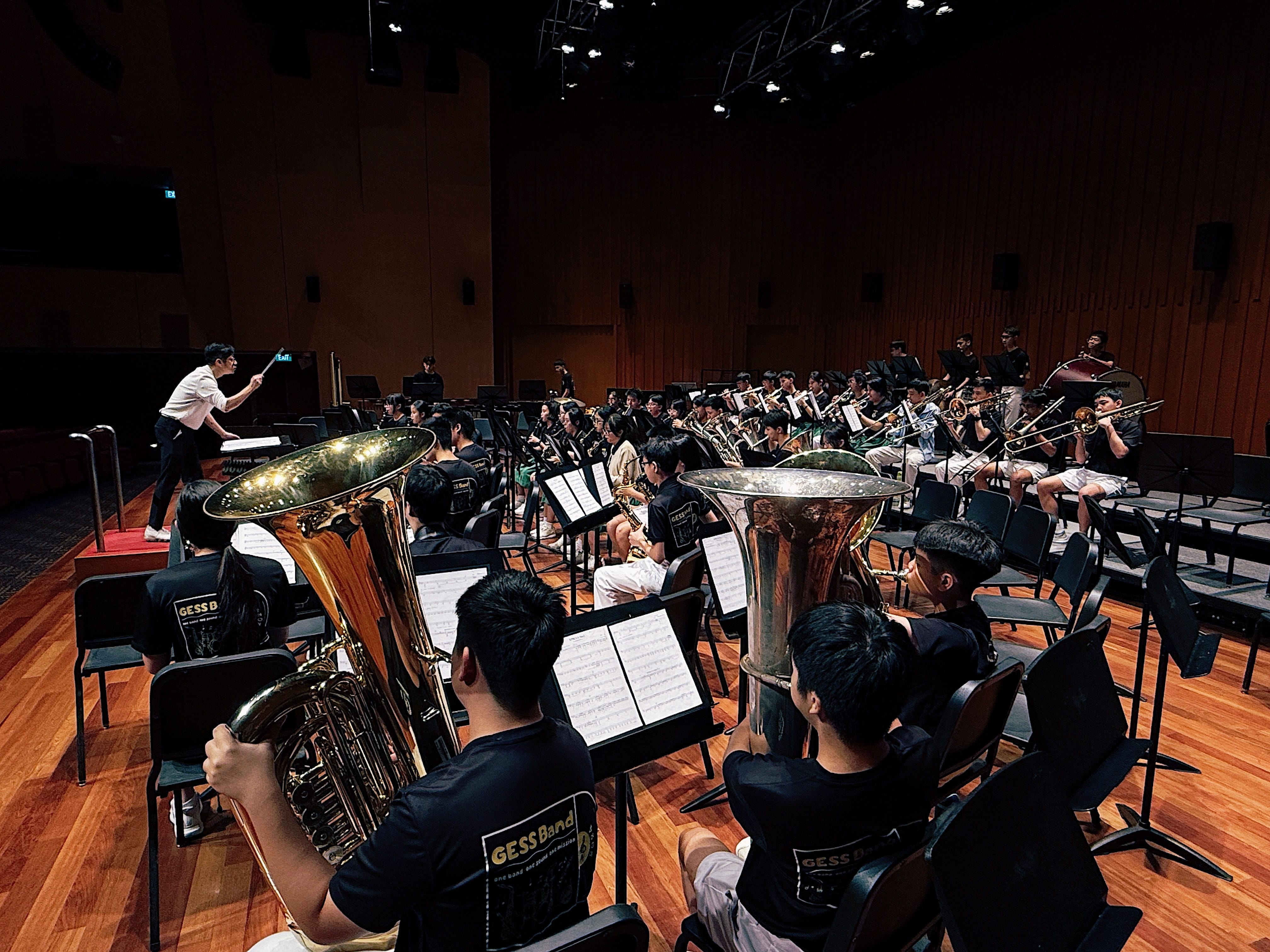 Large youth wind band in black GESS Band shirts rehearsing with tubas and brass instruments, led by a conductor on stage.