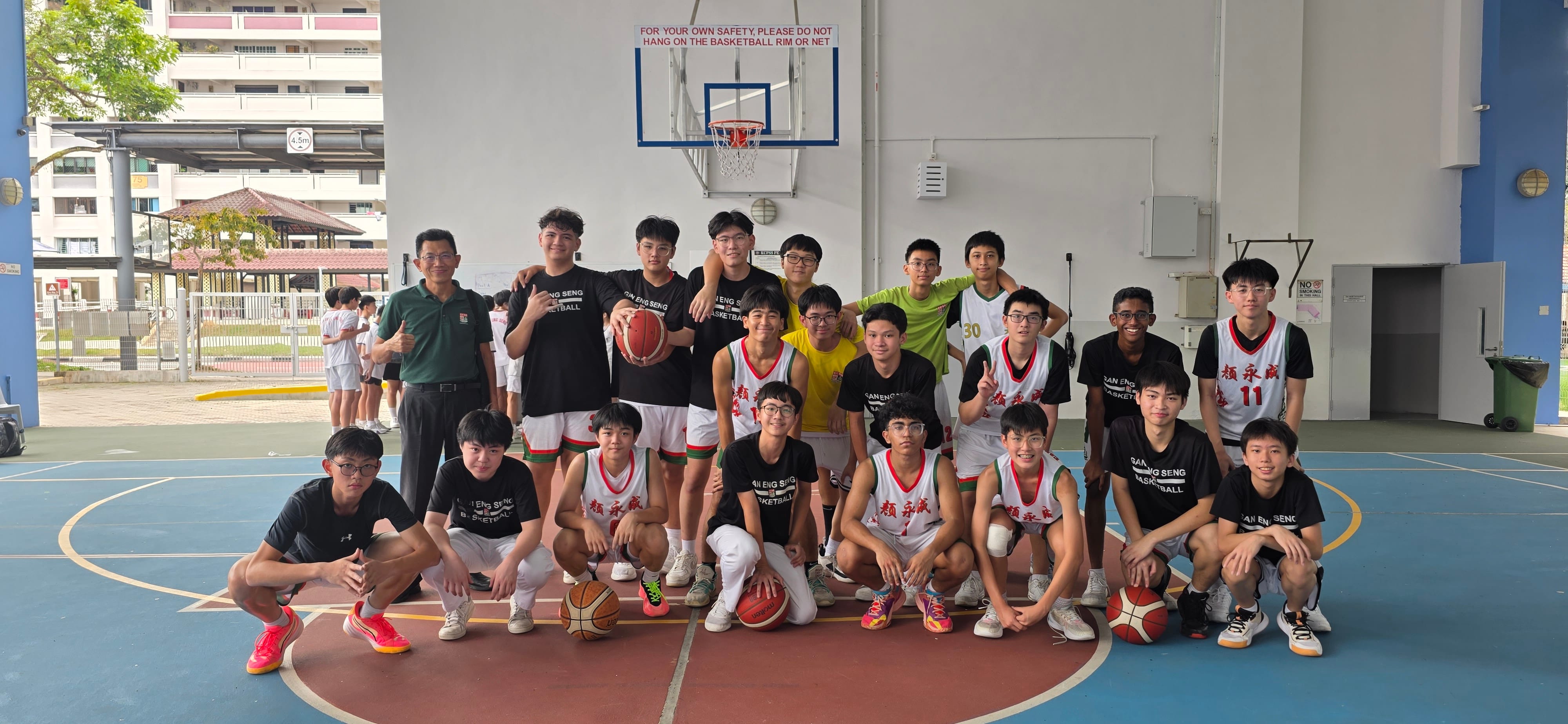 Group of students and a coach pose on an indoor basketball court under a hoop, some holding balls.