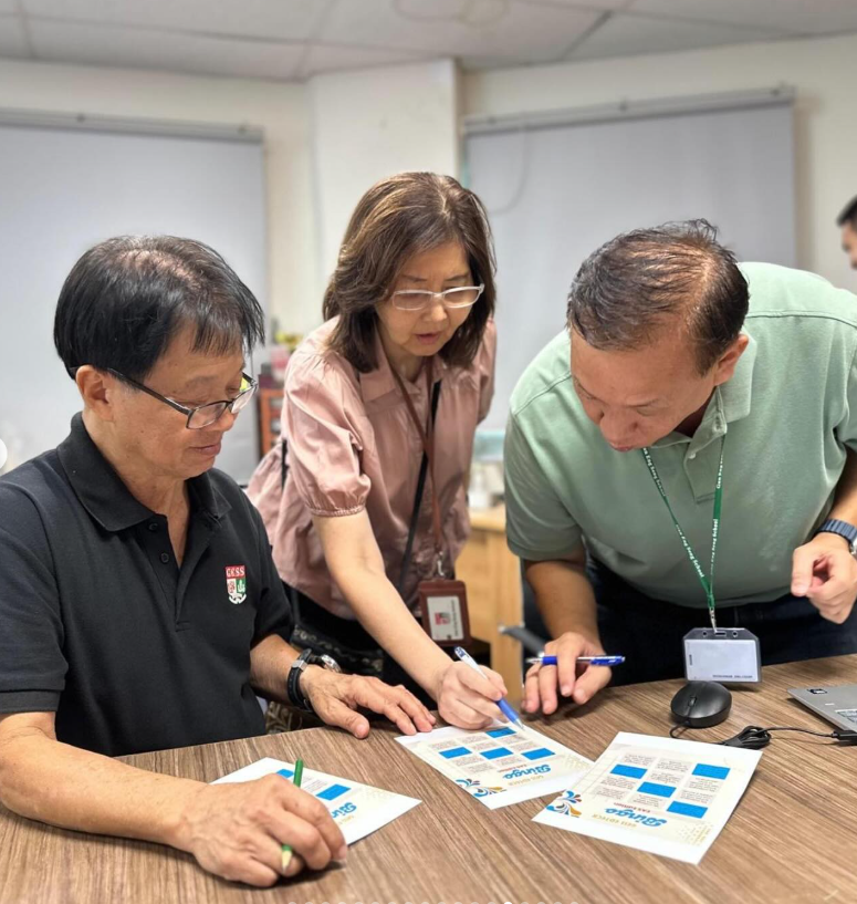 Three people gathered around a table, pointing at printed bingo cards in an office setting.