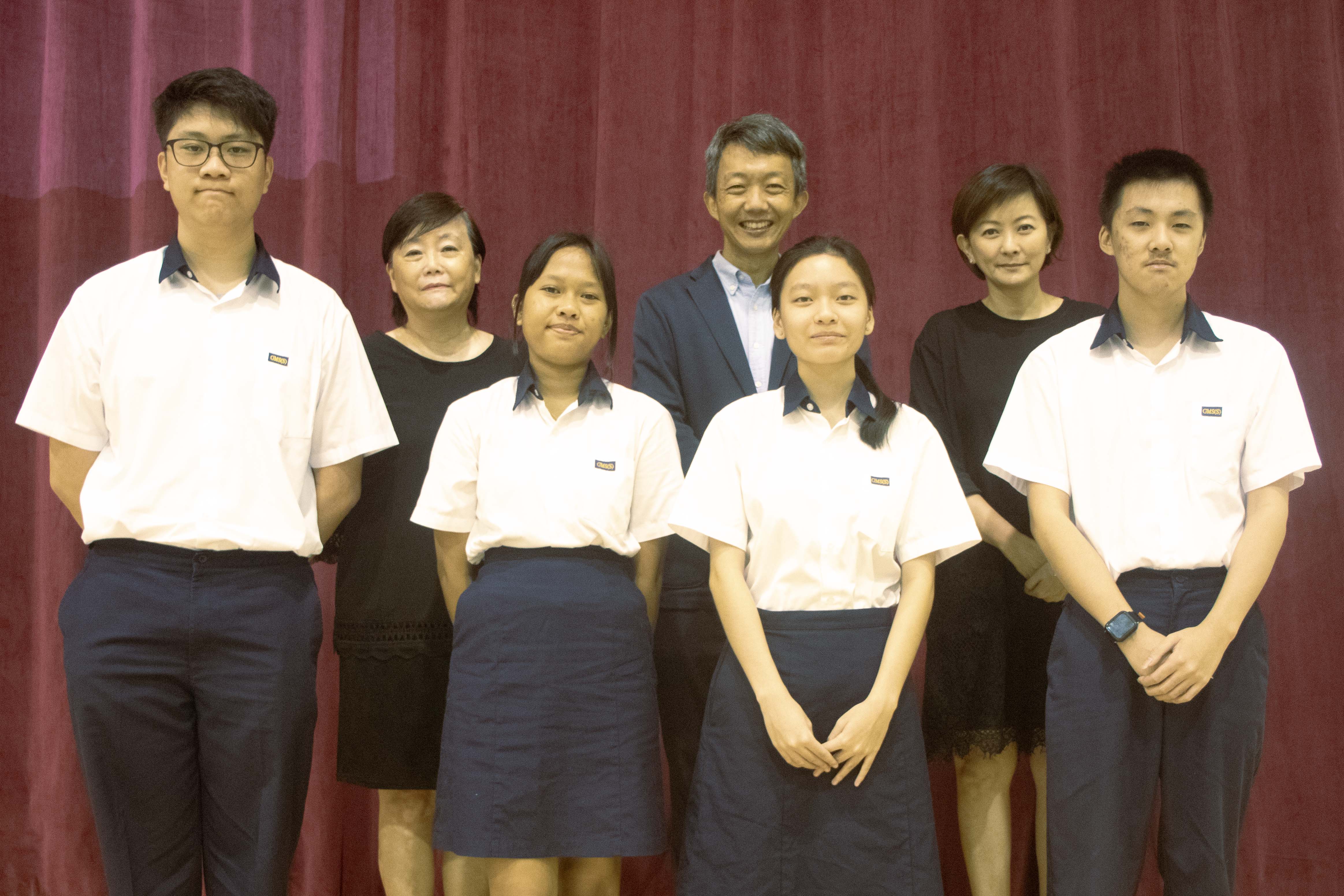 Seven people including four students in white and navy uniforms pose together in front of a red curtain.