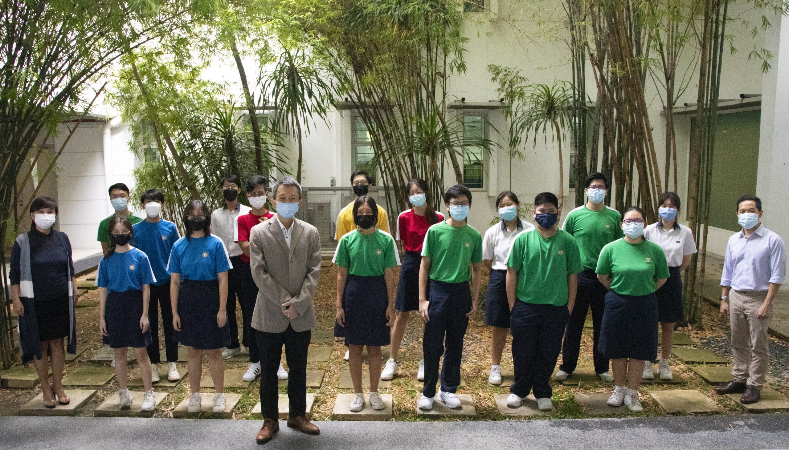 A group of masked students in school uniforms and two adults pose outdoors among bamboo trees near a building.