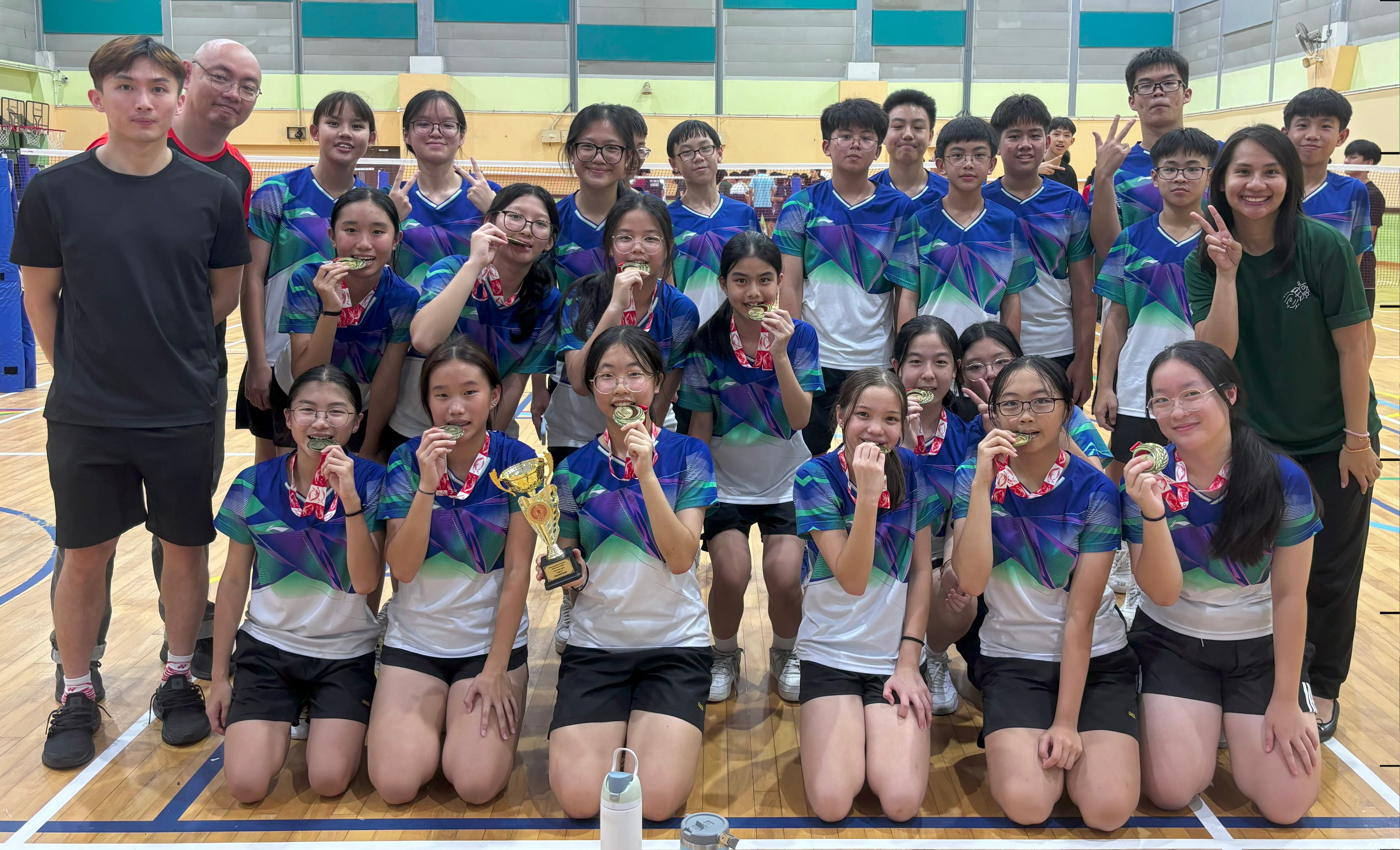 A large group of young athletes in blue jerseys pose in a gym, holding gold medals and a trophy.