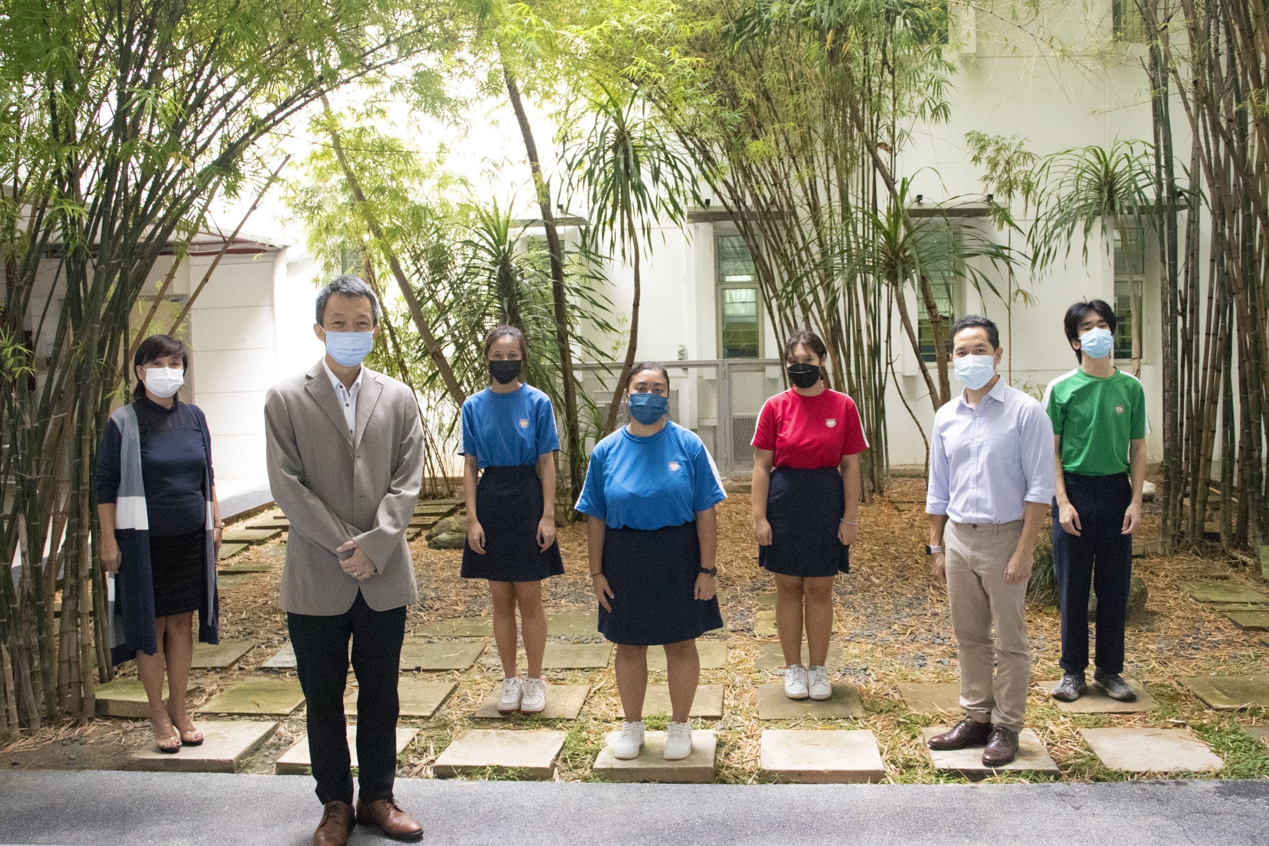 Seven people wearing face masks stand spaced apart in an outdoor courtyard with bamboo trees and a white building.