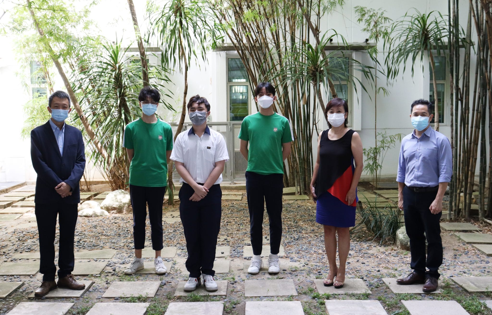 Six people wearing face masks stand outdoors on a stone path surrounded by bamboo and trees near a white building.