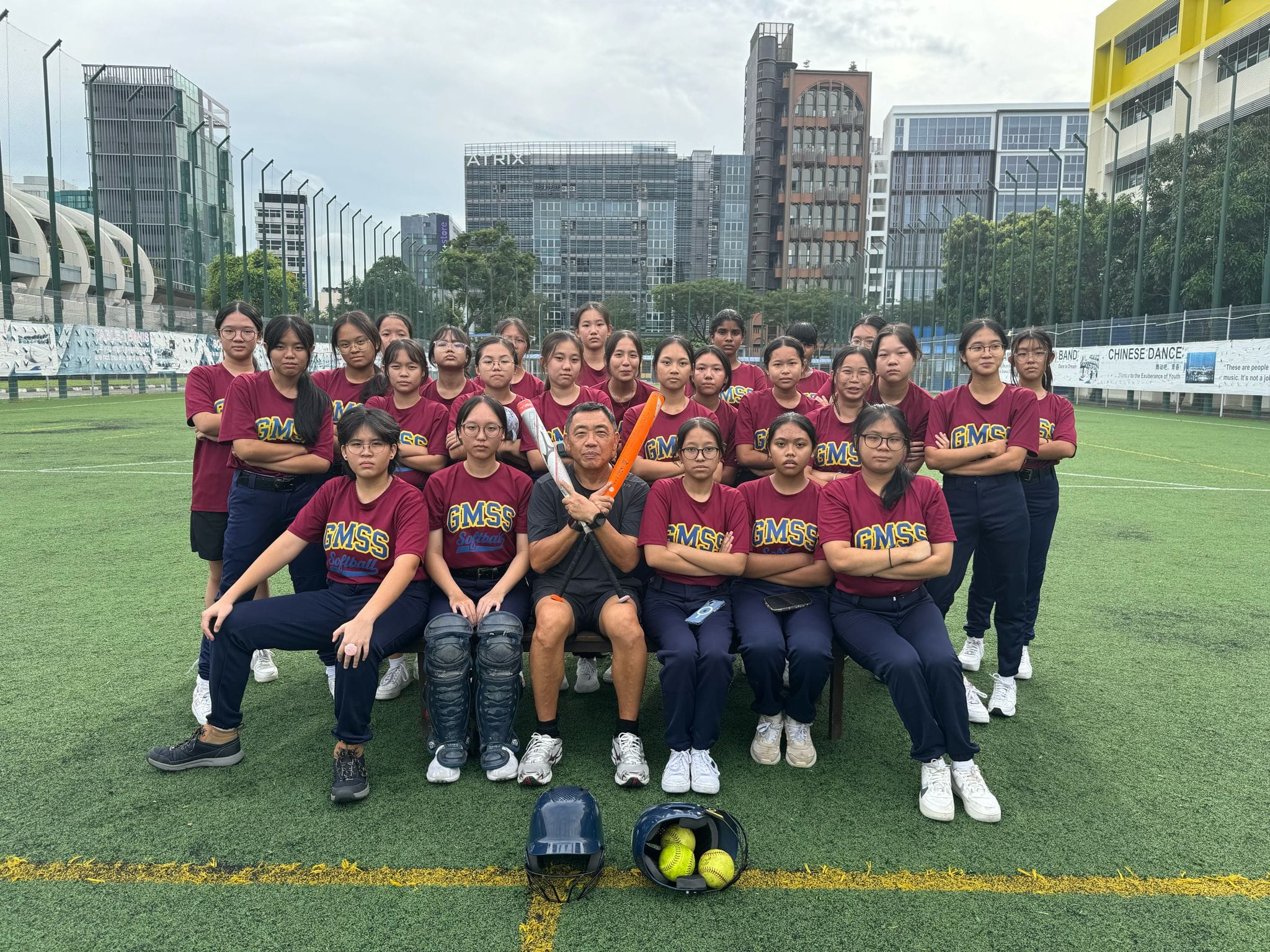 GMSS softball team of girls in maroon shirts posing with a coach holding a bat on a green field.