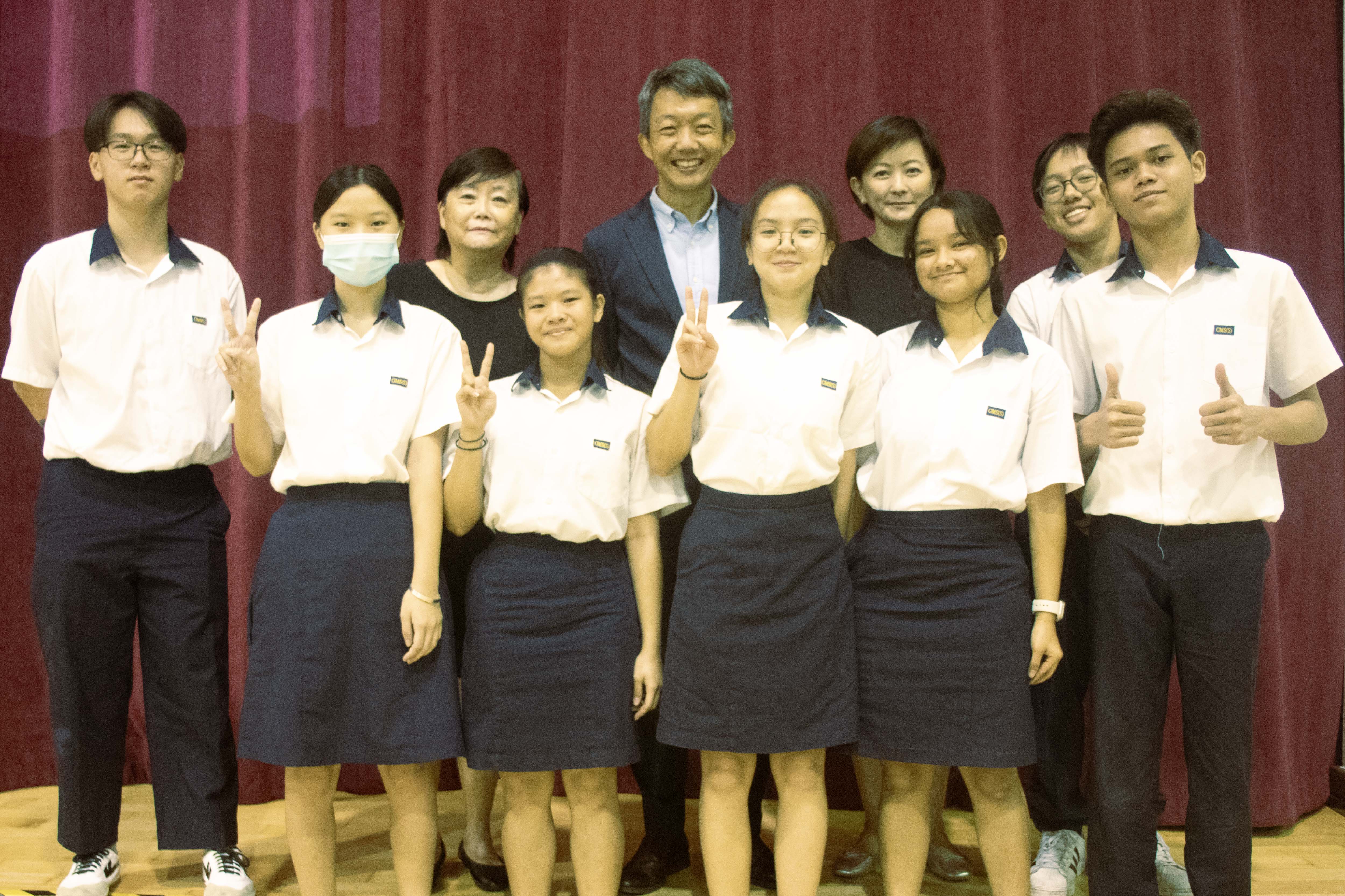 Group of students in white and navy uniforms posing with two adults in front of a red curtain.