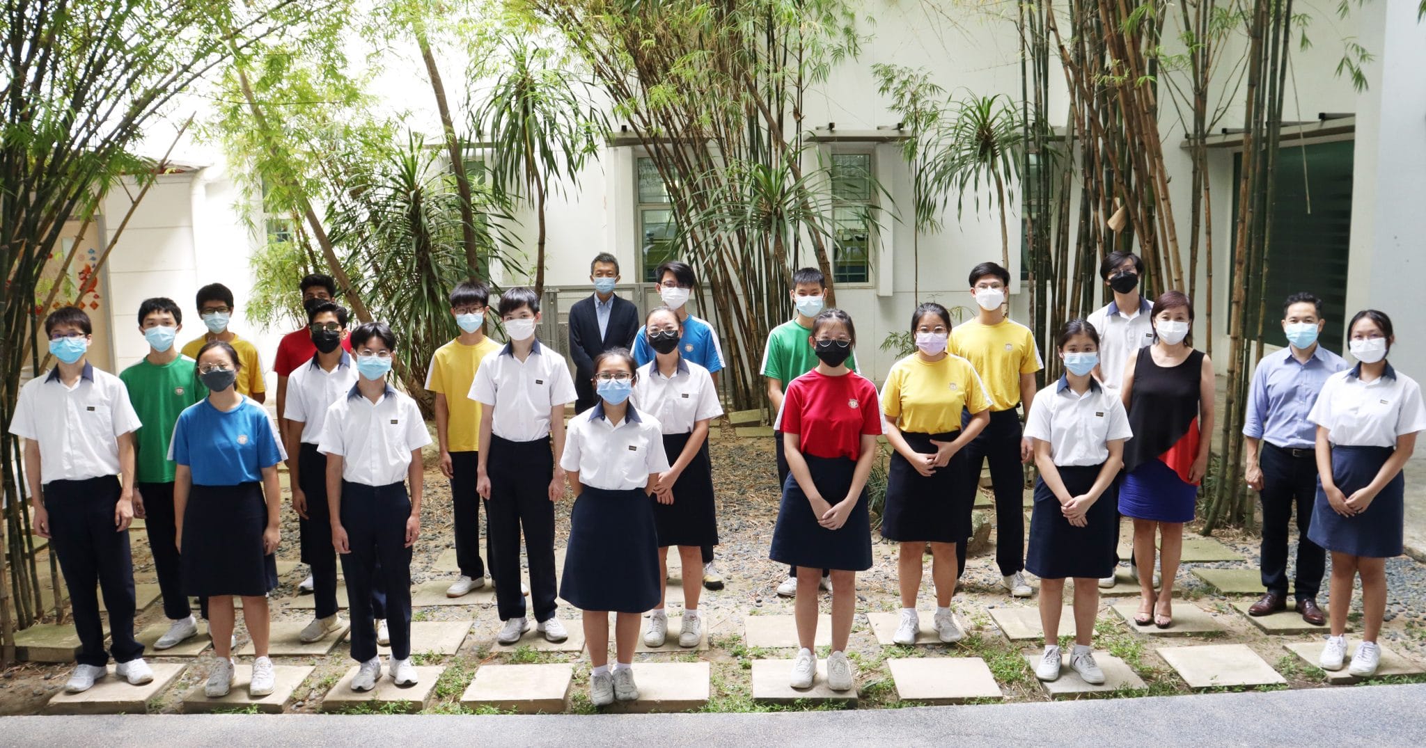 A group of students and adults wearing face masks pose outdoors near bamboo trees beside a white building.