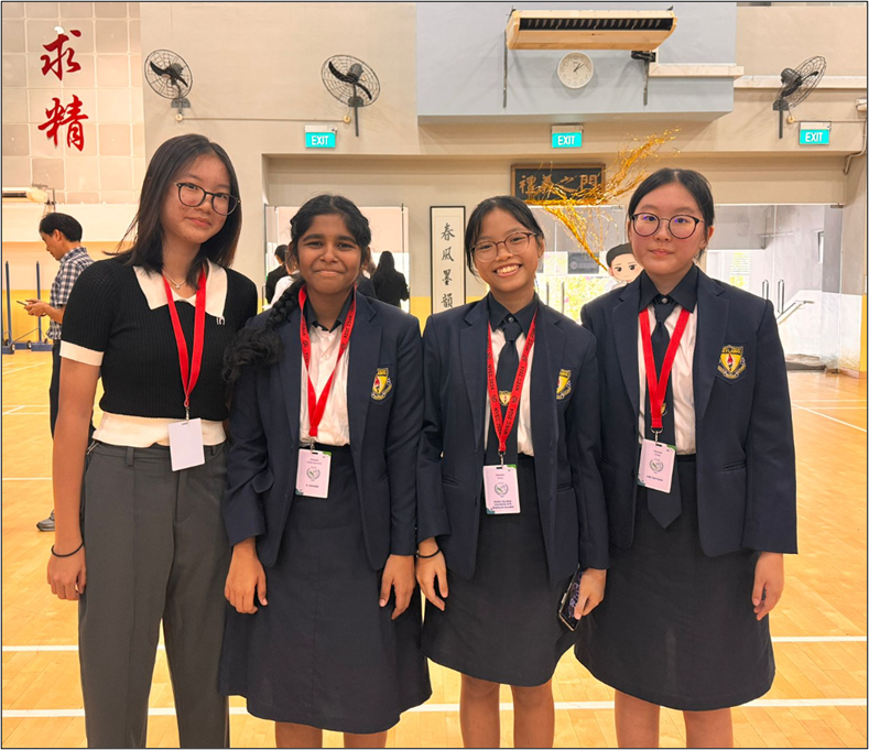 Four girls in school uniforms with red lanyards pose together in a hall with Chinese calligraphy on the walls.