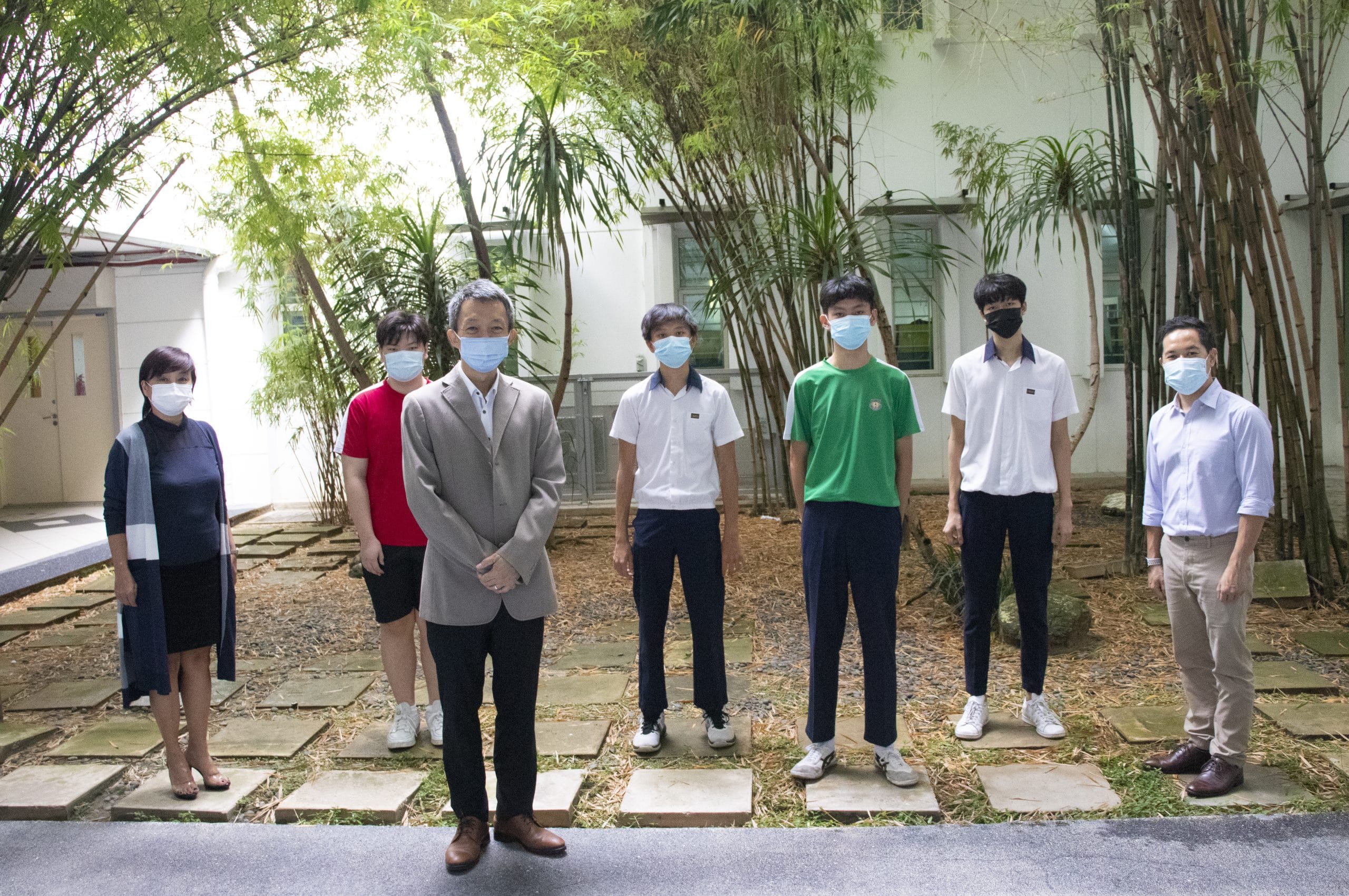 Seven people wearing face masks stand spaced apart on stone pavers in a courtyard with bamboo trees.