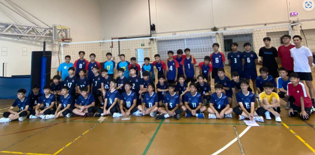 A large group of young male volleyball players posing together in an indoor gymnasium with a net behind them.