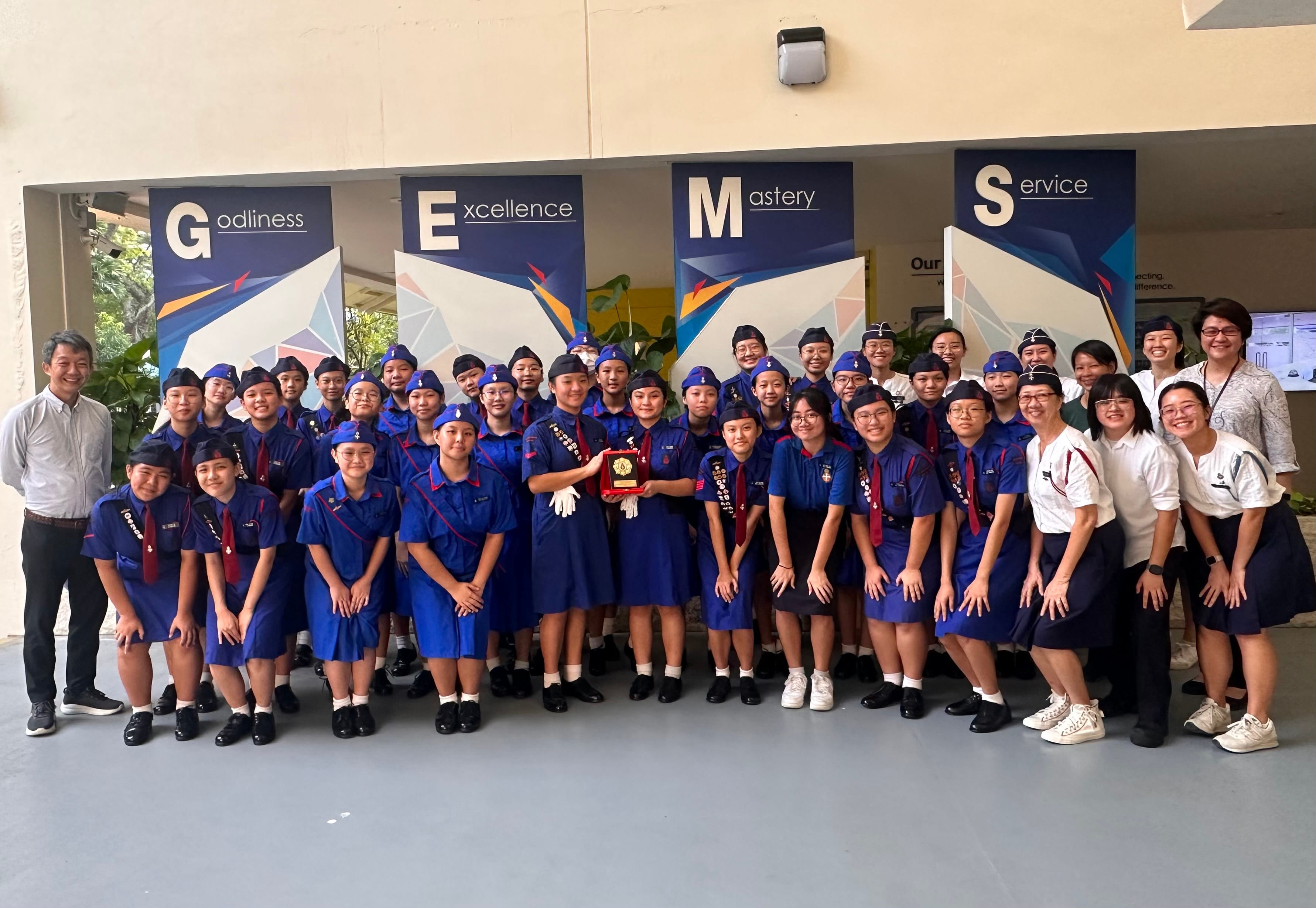 A large group of students in blue uniforms and caps pose with a trophy in front of GEMS banners.