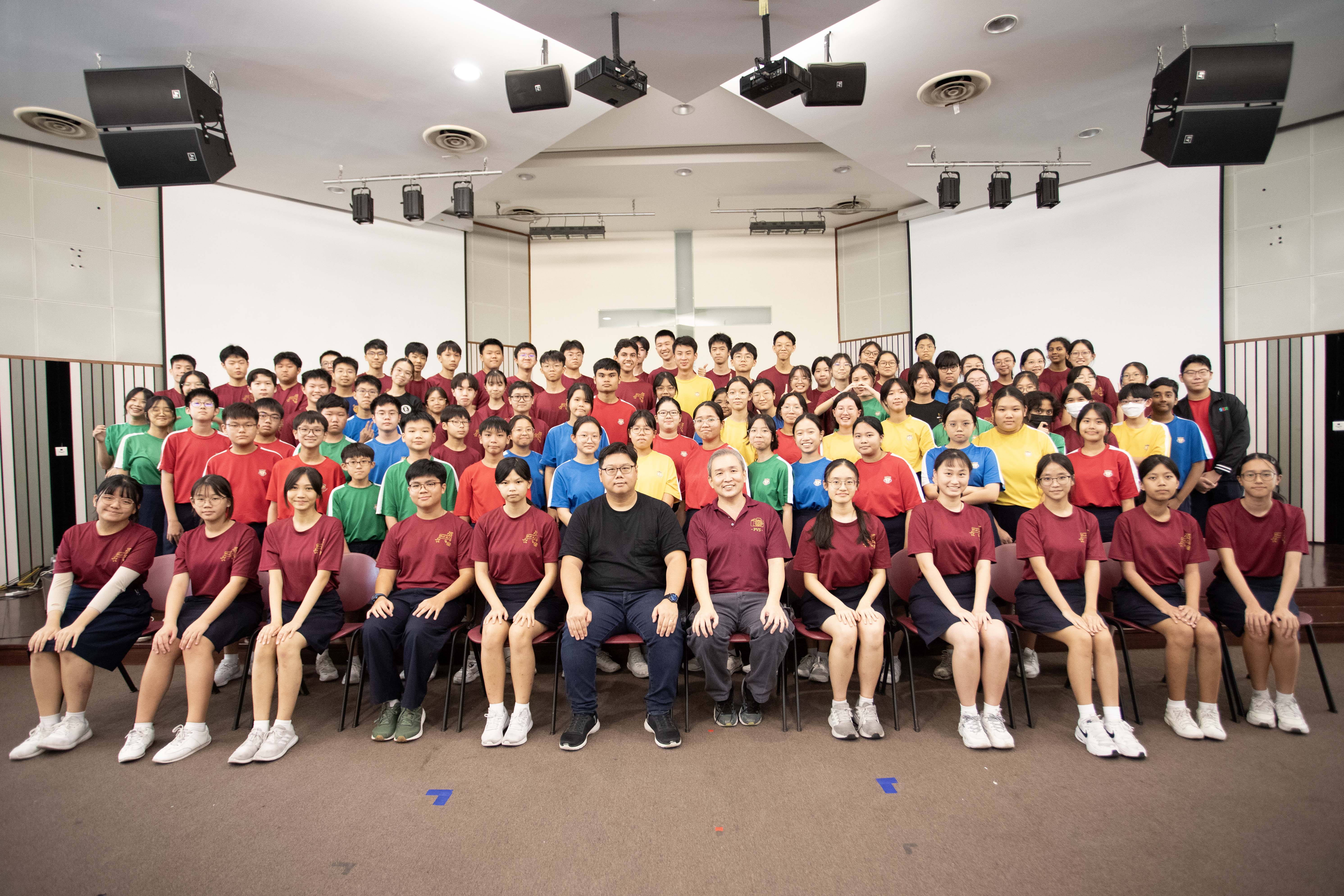Large group of students in colorful uniforms posing with two adults in an indoor auditorium for a group photo.
