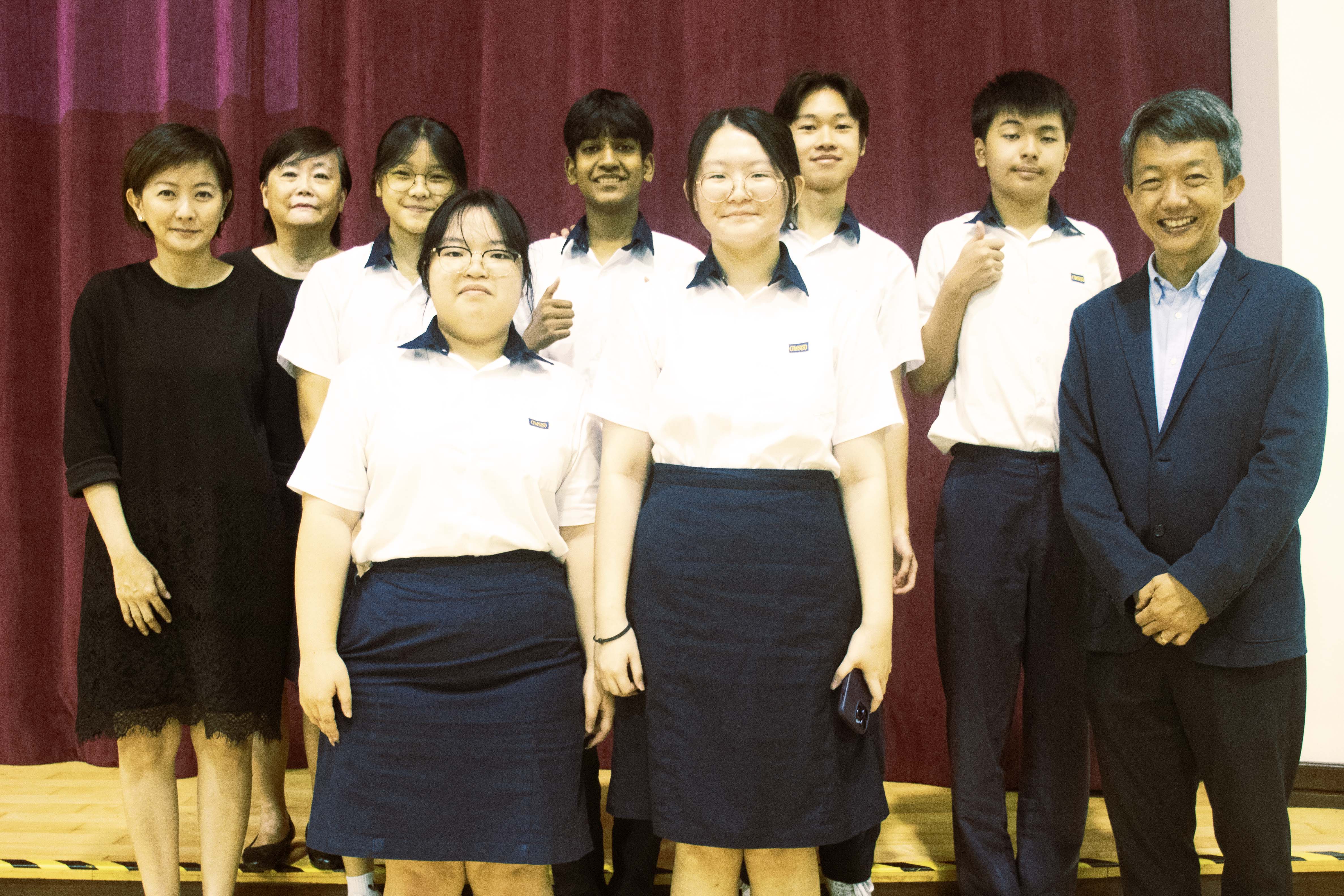 Nine people pose for a group photo on a wooden stage in front of a red curtain, students in white uniforms flanked by adults.