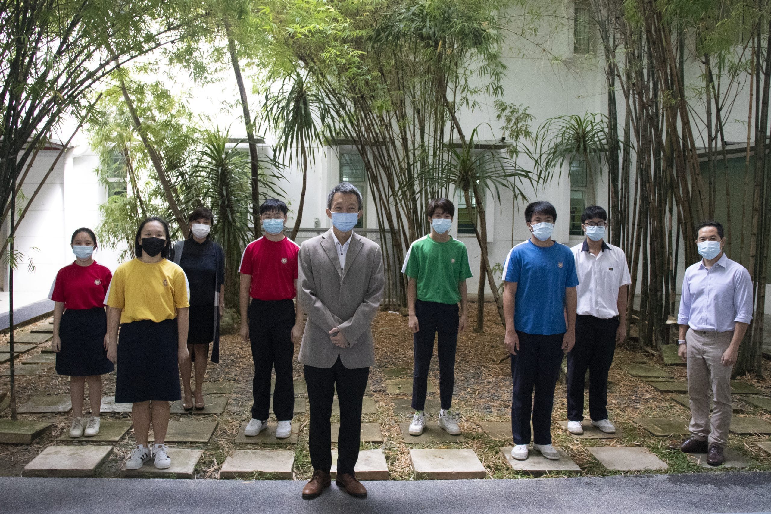 Nine people wearing face masks stand spaced apart outdoors in a courtyard with bamboo trees and a white building behind them.